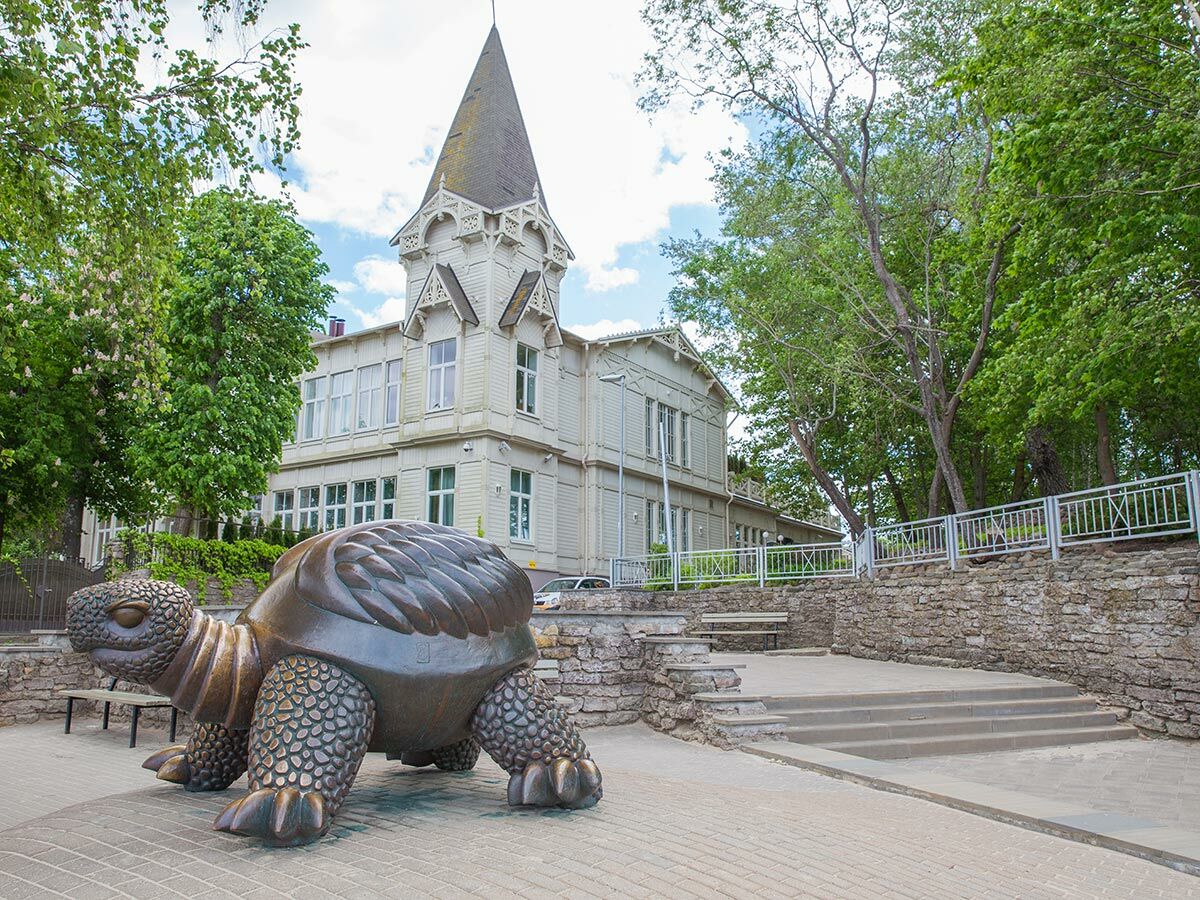 Große Bronzestatue einer Schildkröte auf gepflastertem Platz, dahinter weiße historische Villa mit Turm und Bäumen.