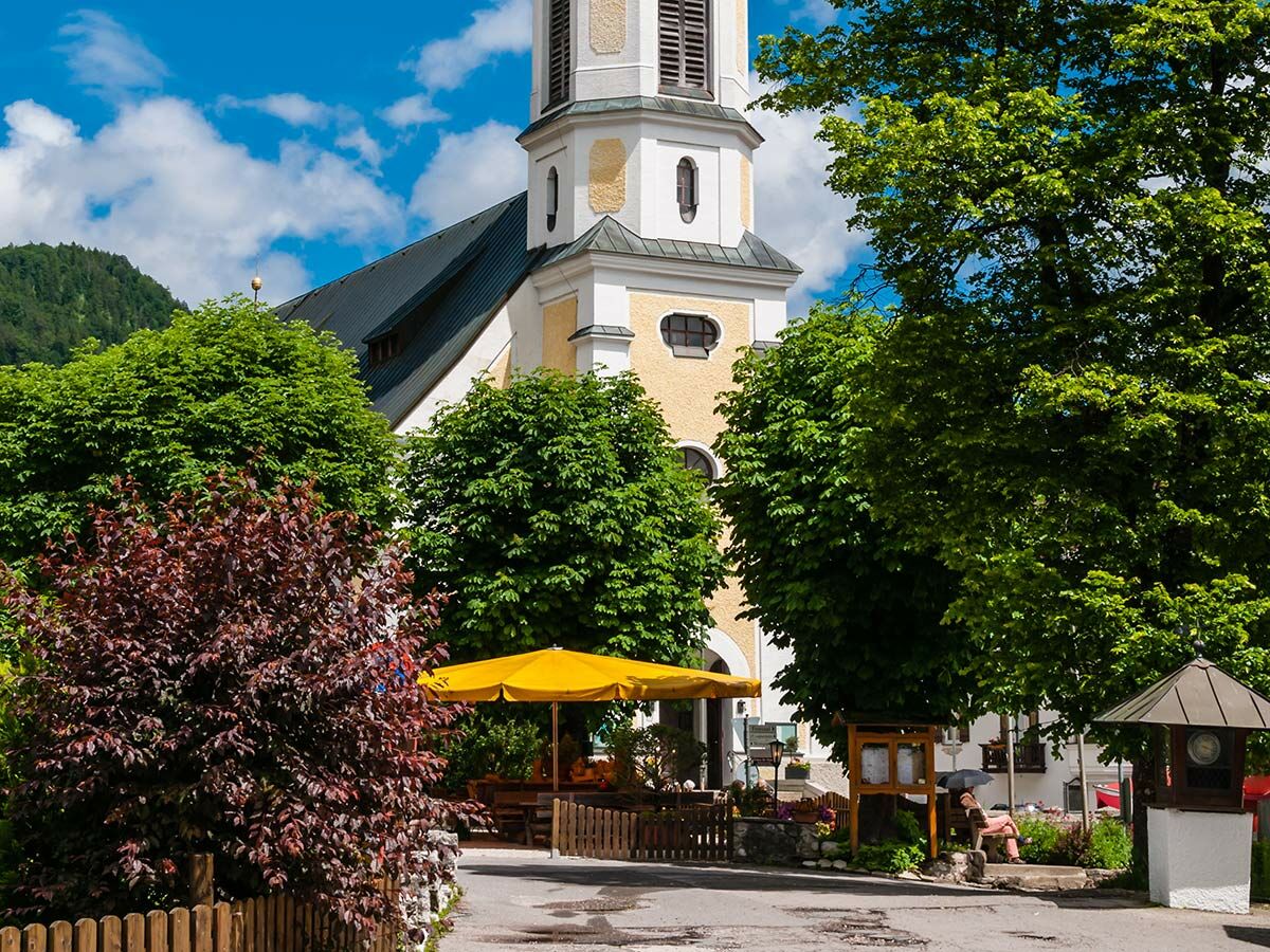 Idyllische Dorfkirche mit hohem Turm und gelb-weißer Fassade, umgeben von grünen Bäumen, rotem Busch bei sonnigem Wetter in den bayerischen Alpen.