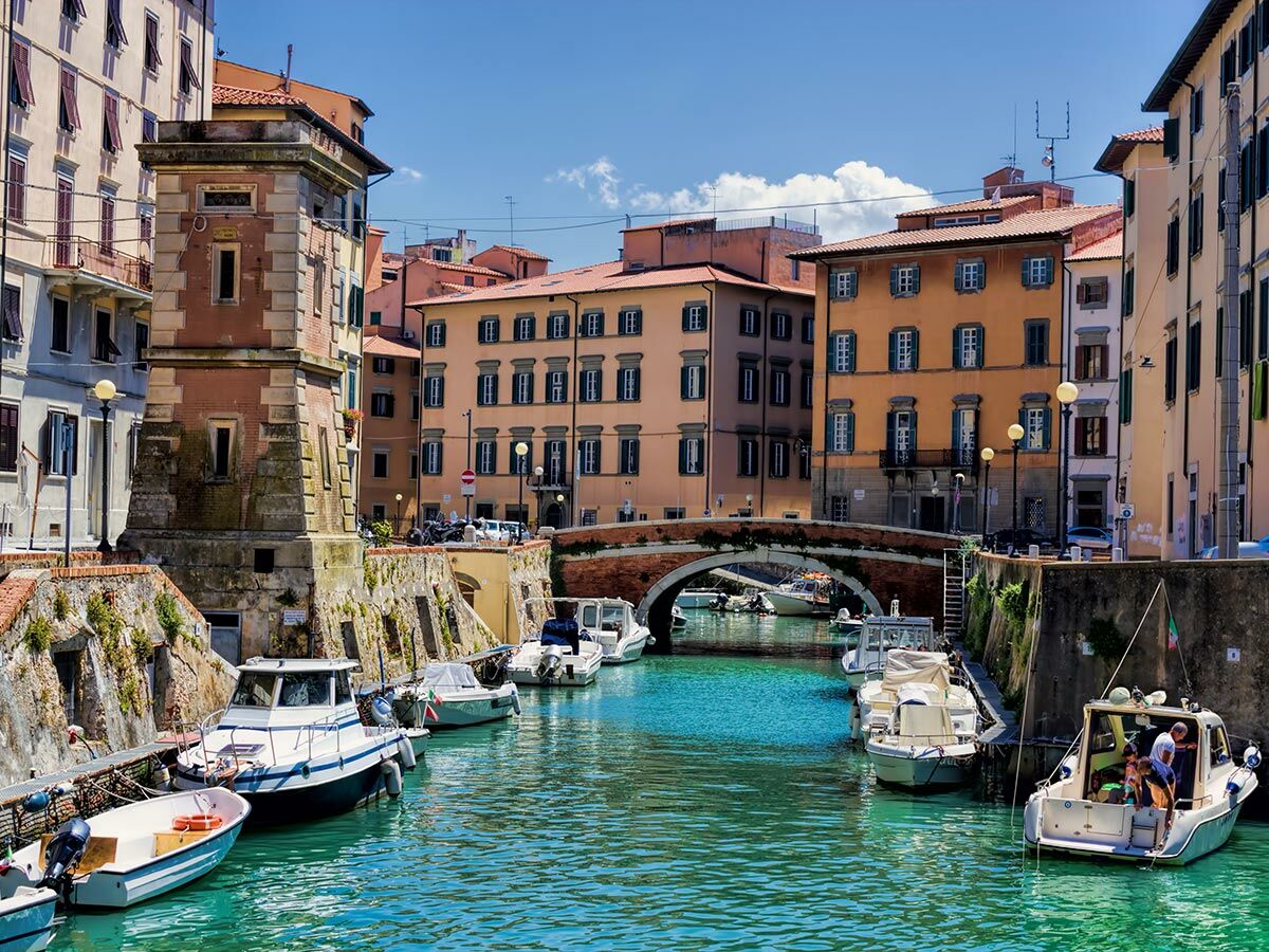Historische italienische Kanalstadt mit bunten Gebäuden, Brücke und Booten auf türkisfarbenem Wasser an sonnigem Tag.