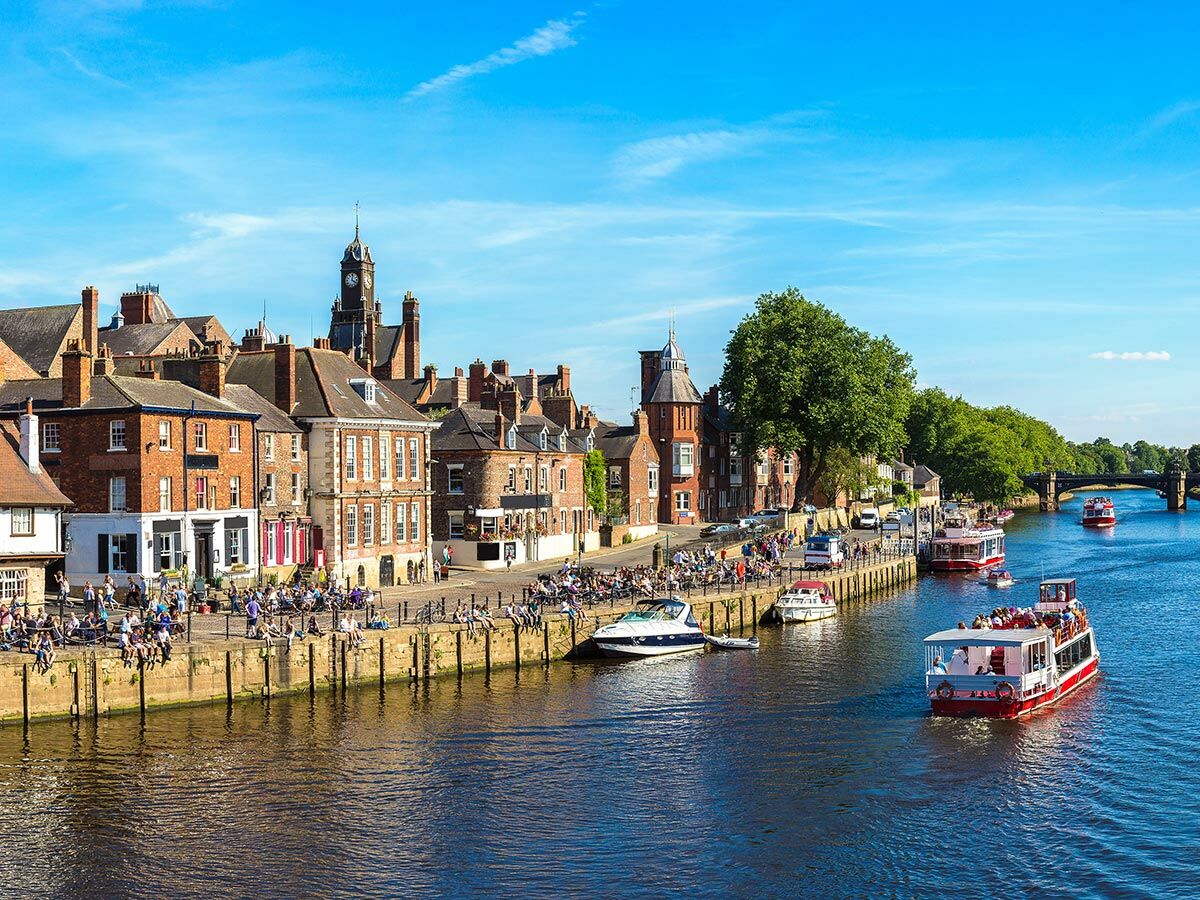 Historische Gebäude säumen den Fluss Ouse in York, Menschen entspannen am Ufer, Touristenboote fahren bei blauem Himmel.