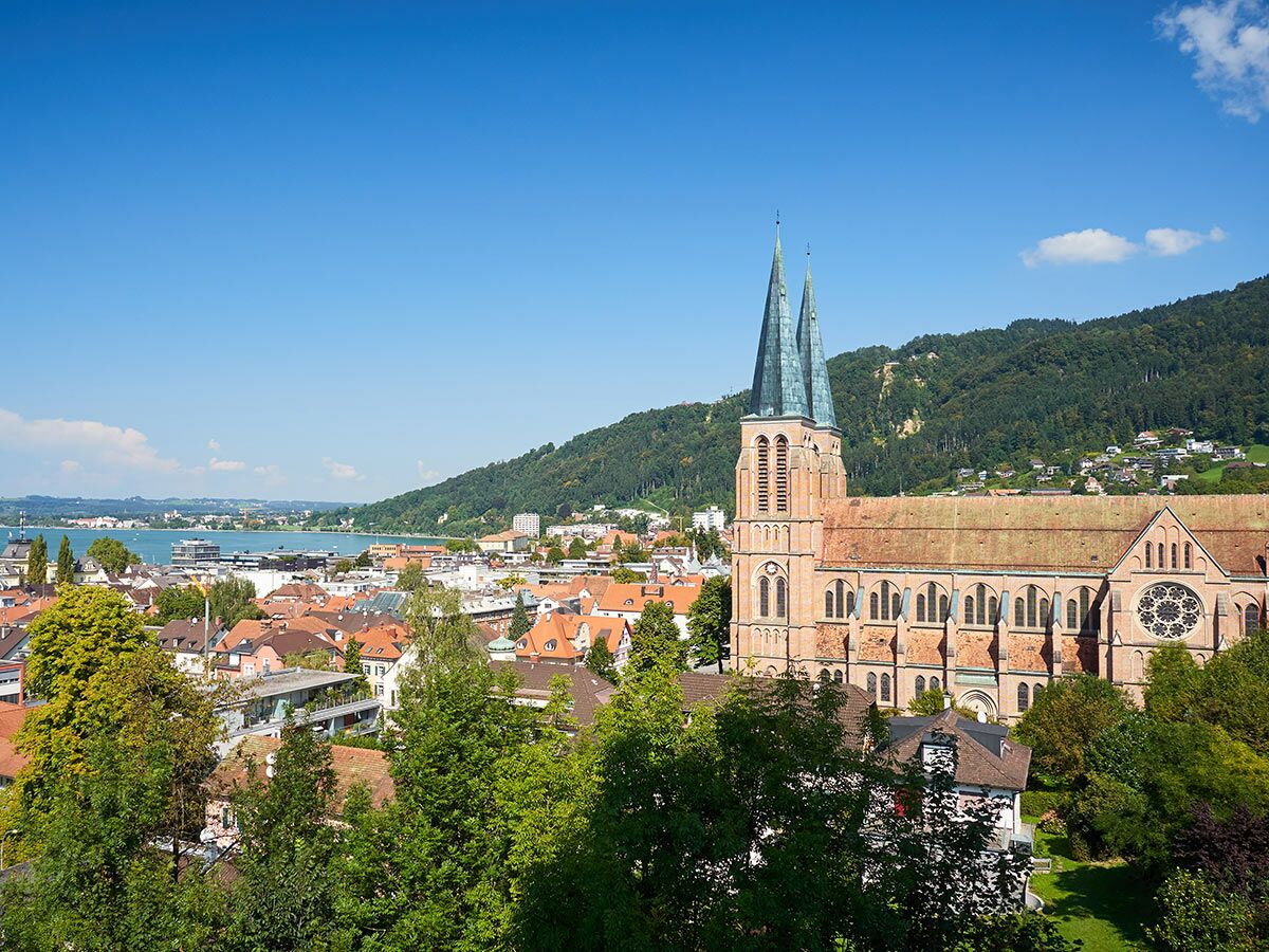 Luftaufnahme von Bregenz, Österreich, mit der Herz-Jesu-Kirche, roten Dächern und dem Bodensee unter blauem Himmel.
