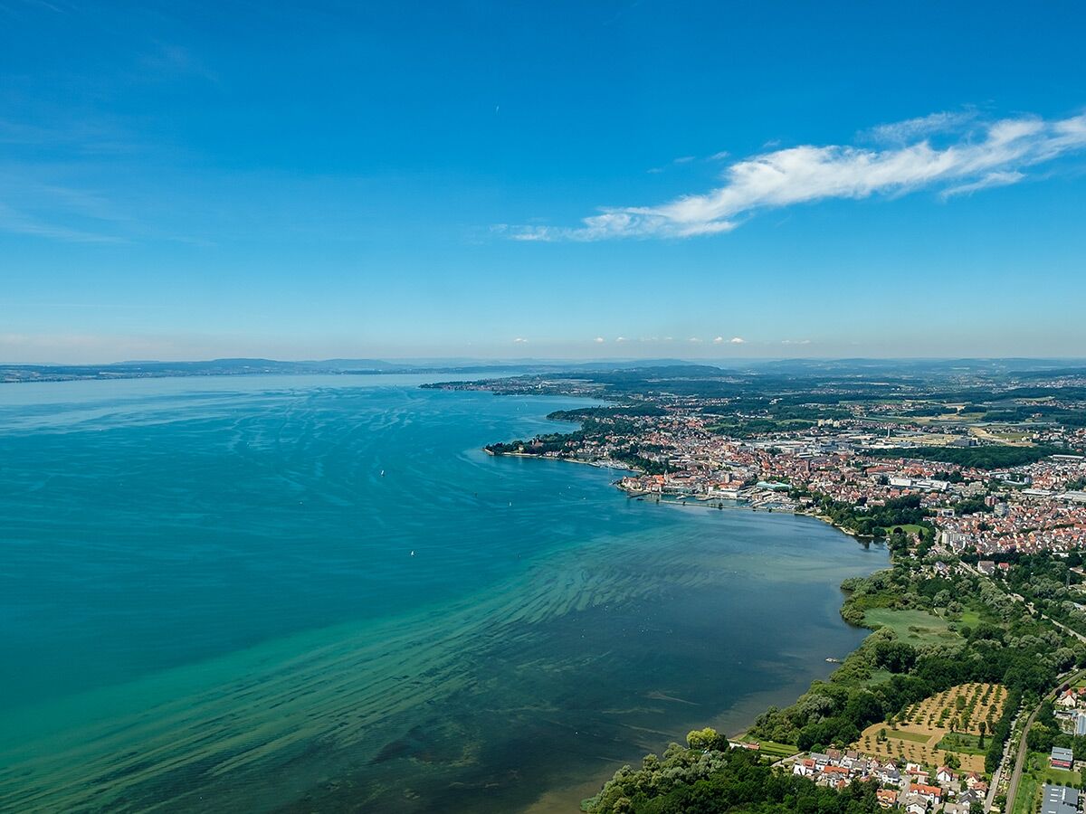 Luftaufnahme von Radolfzell am Bodensee mit türkisfarbenem Wasser, Stadtbild, Booten und blauem Himmel.