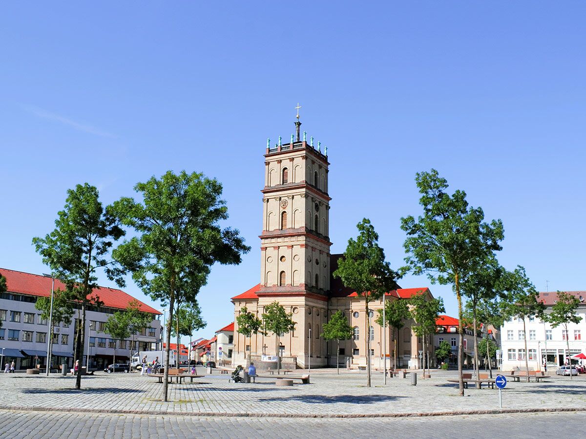 Ansicht eines historischen Kirchturms und eines belebten Marktplatzes mit Bäumen und Menschen unter blauem Himmel.
