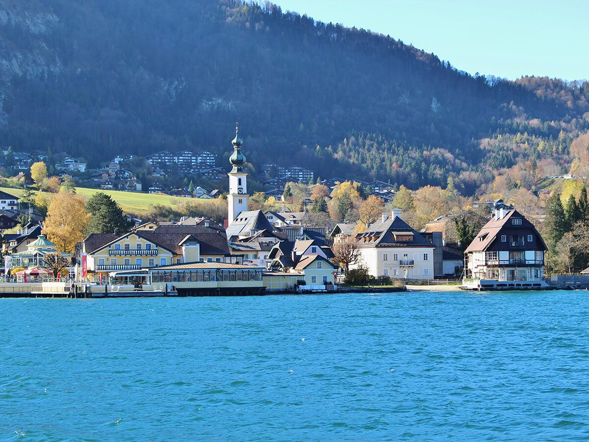 Idyllisches Dorf mit Kirche, bunten Häusern am blauen See, umgeben von herbstlichen Alpenbergen unter klarem Himmel.