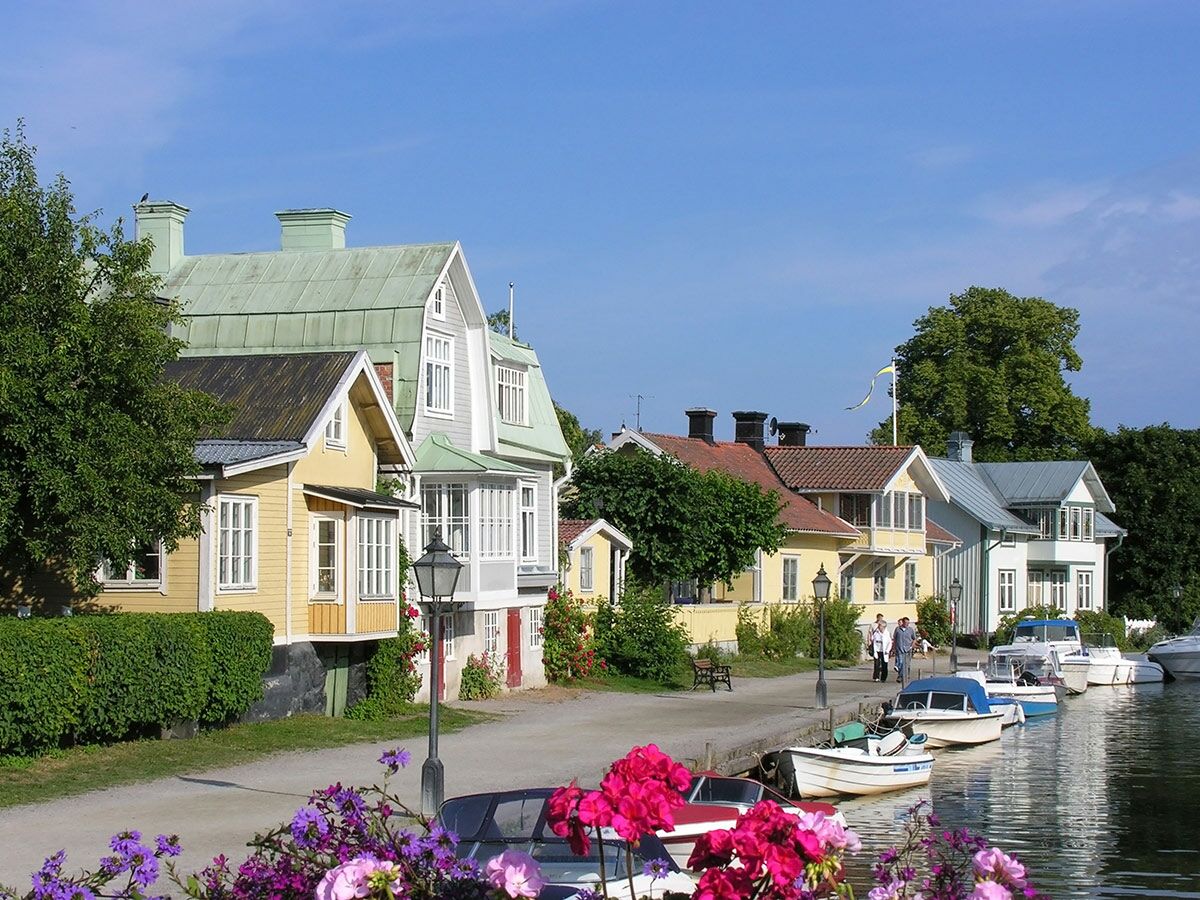 Idyllisches schwedisches Dorf am Kanal mit bunten Holzhäusern, vielen Booten und Menschen unter blauem Sommerhimmel.
