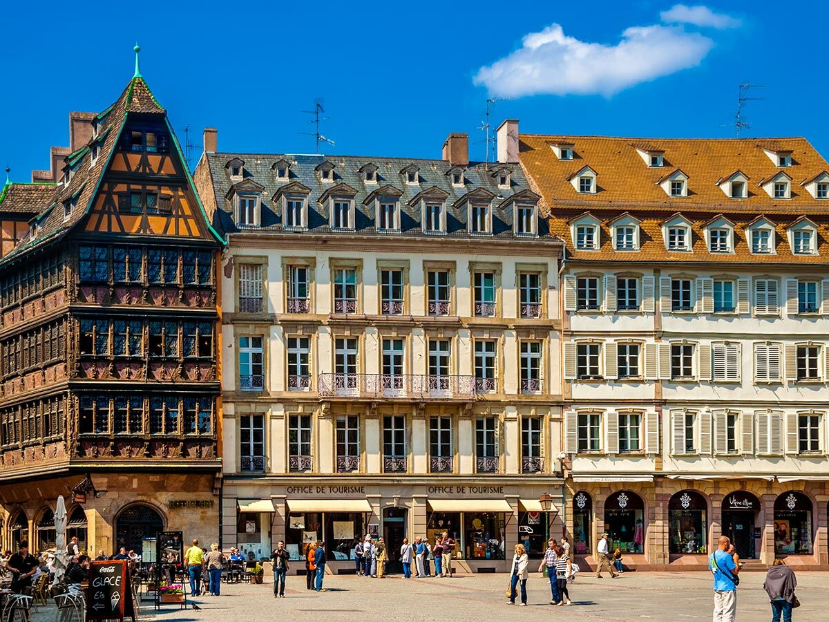 Historische Gebäude in Strasbourg am Münsterplatz mit Fachwerkhäusern, Touristenbüro und Menschen bei sonnigem Wetter.