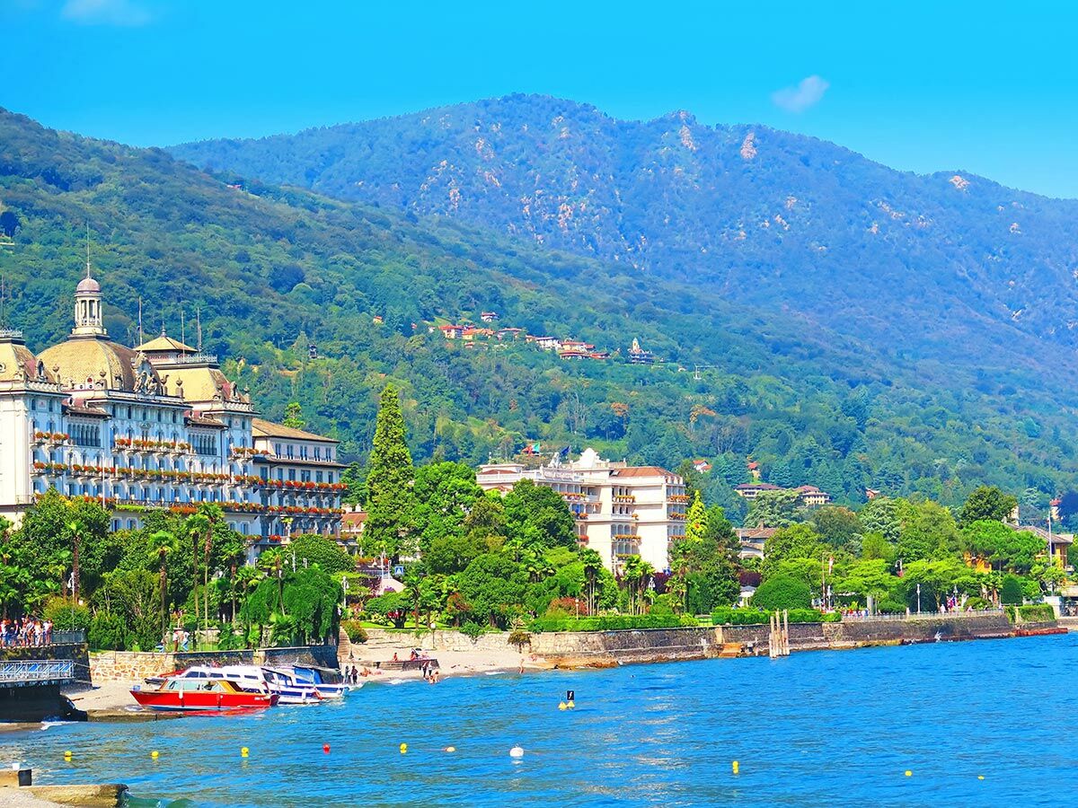 Elegantes Seehotel an einem Bergsee mit blauen Booten, grünen Palmen und einer belebten Uferpromenade.