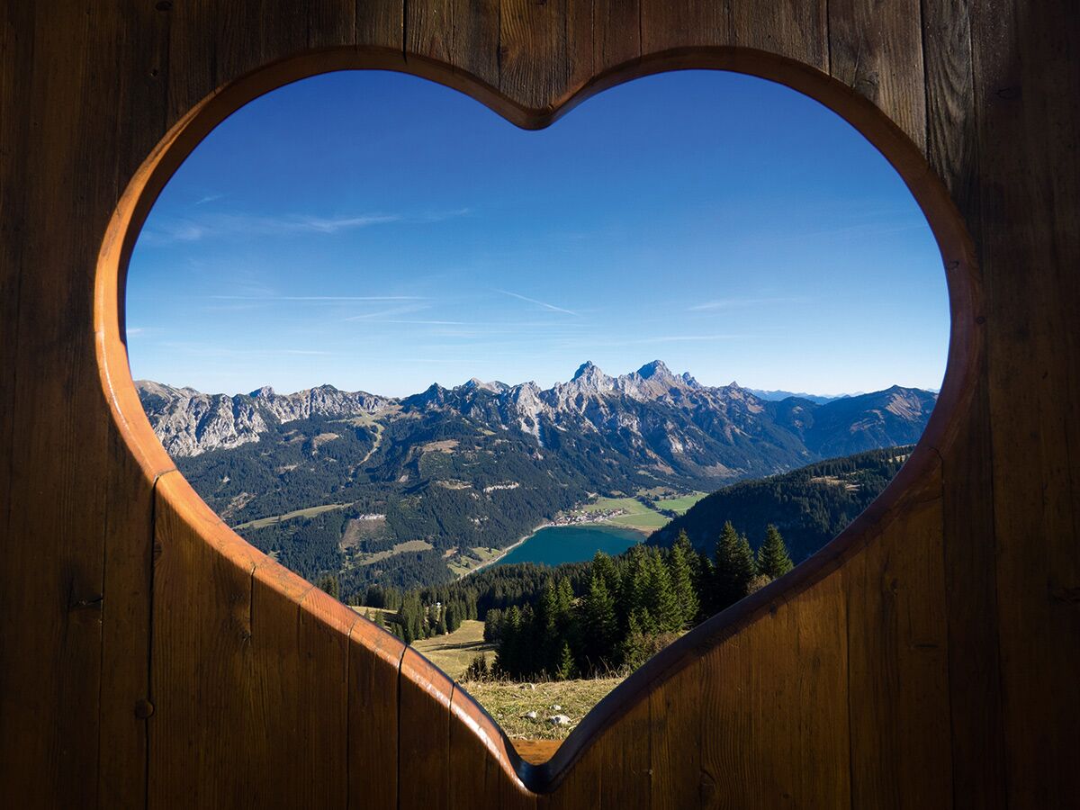 Malerisches Bergpanorama mit See und Wäldern, gerahmt von einem herzförmigen Holzausschnitt vor blauem Himmel.