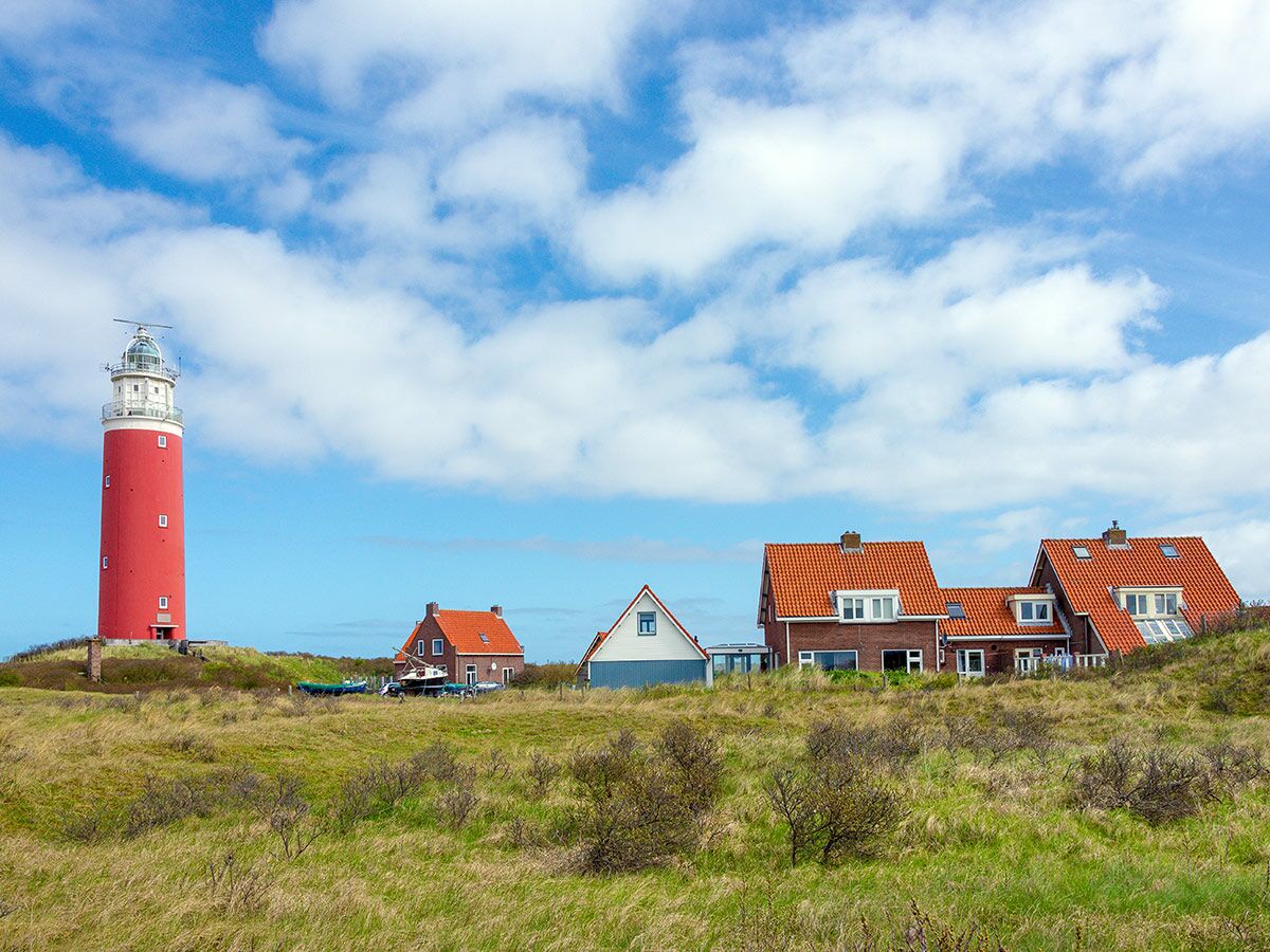 Roter Leuchtturm auf grüner Düne mit typischen Häusern und blauem Himmel an der Küste von Texel, Niederlande.