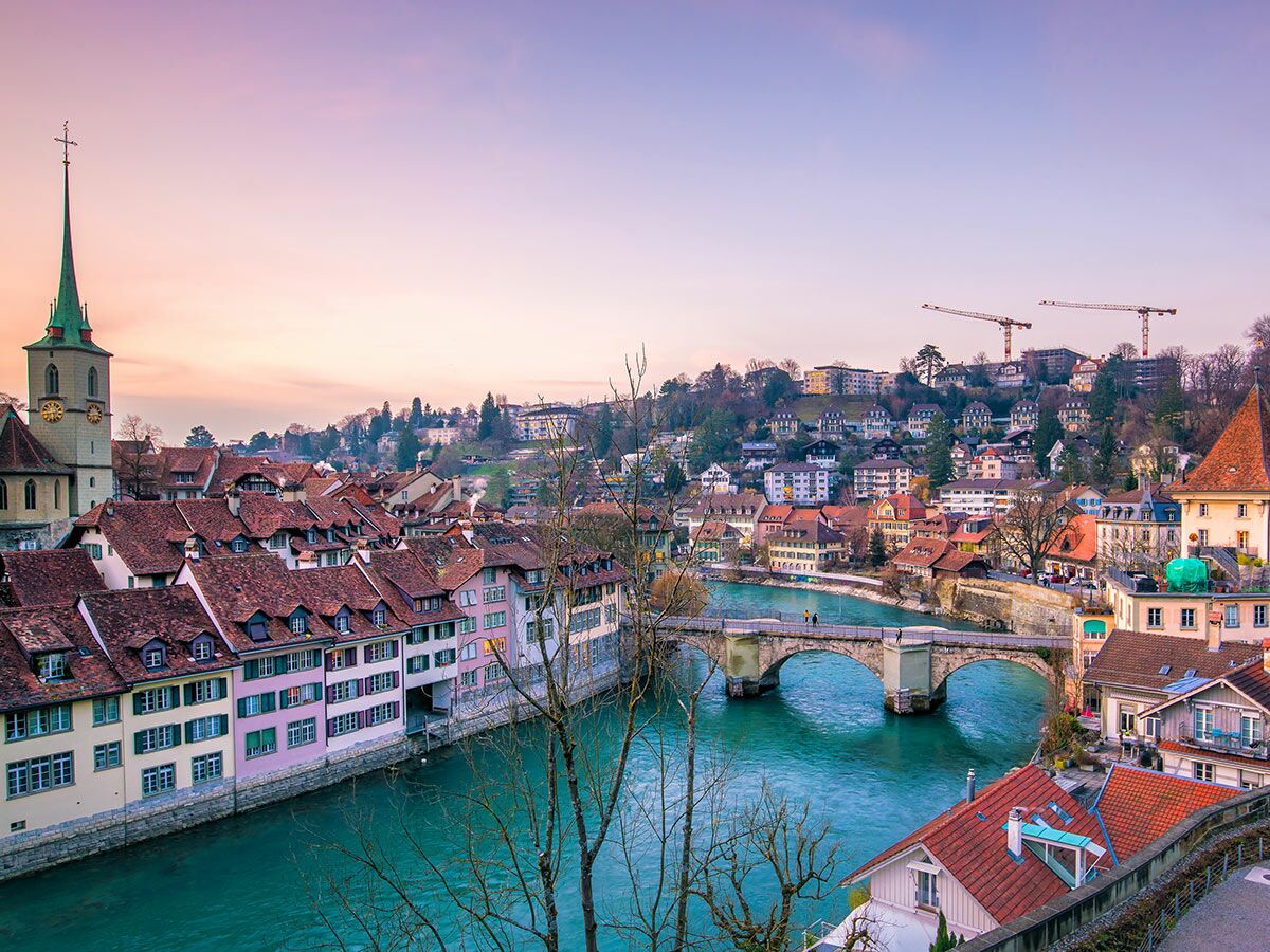Panoramablick auf die Berner Altstadt am Fluss Aare bei Sonnenuntergang mit historischer Brücke, bunten Häusern und Kirchturm.