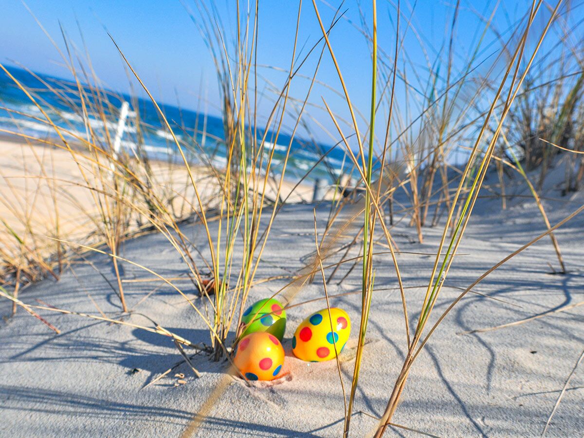 Drei bunte, gepunktete Ostereier versteckt im Sand von Dünen am Strand, mit Meer und blauem Himmel im Hintergrund.