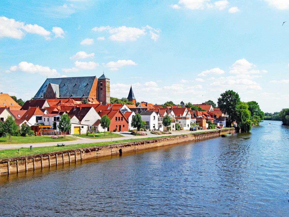 Panorama einer historischen Stadt mit roten Dächern am Fluss, dominiert von einer großen Kirche unter blauem Himmel.