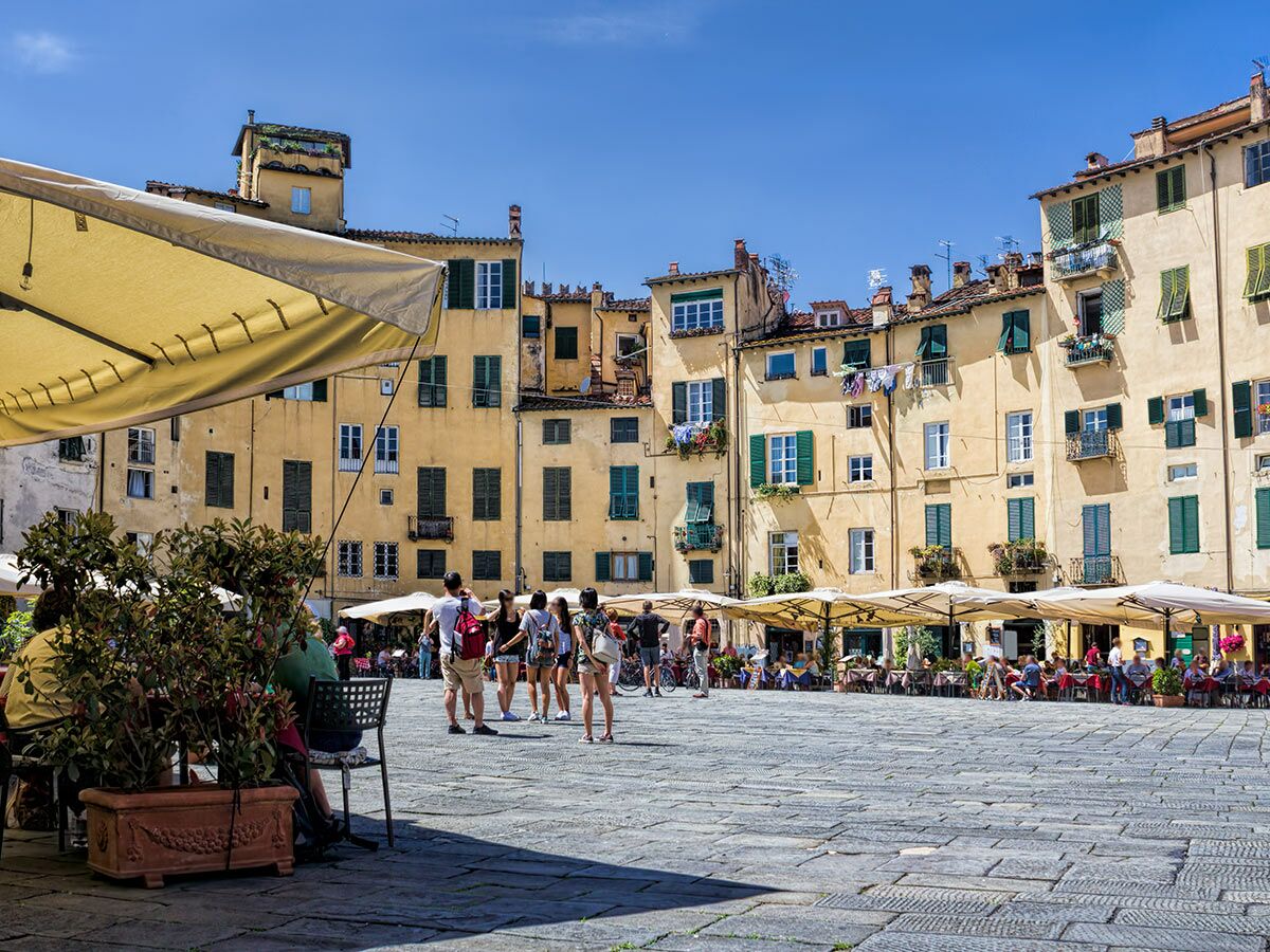 Belebtes Piazza dell'Anfiteatro in Lucca, Italien mit gelben Gebäuden, grünen Fensterläden, Cafés und Menschen bei Sonnenschein.