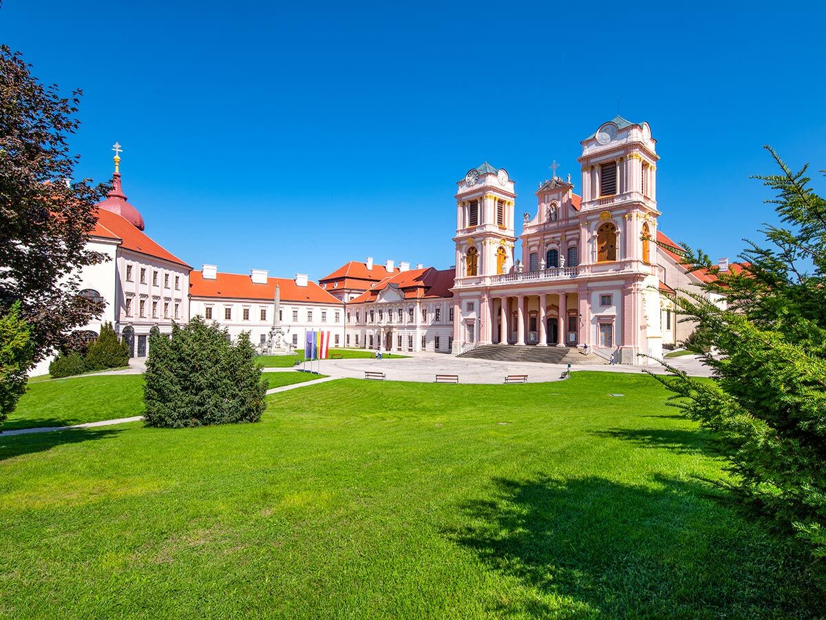 Barocke Stiftskirche Vorau in Österreich mit rosa Fassade, weißen Türmen und grüner Parkanlage unter blauem Himmel.