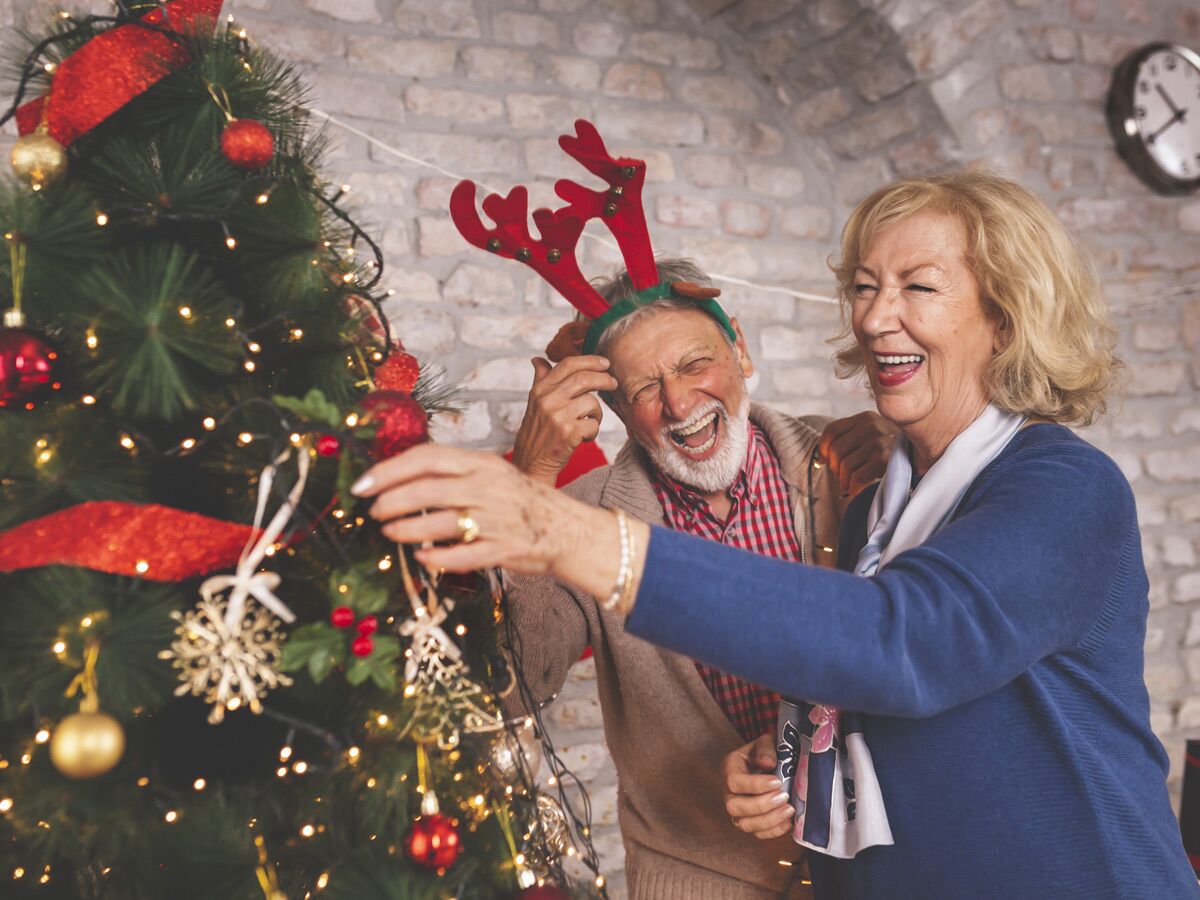 Glückliches Seniorenpaar lacht und schmückt einen Weihnachtsbaum mit Lichtern und roten Kugeln; Mann trägt Rentiergeweih.