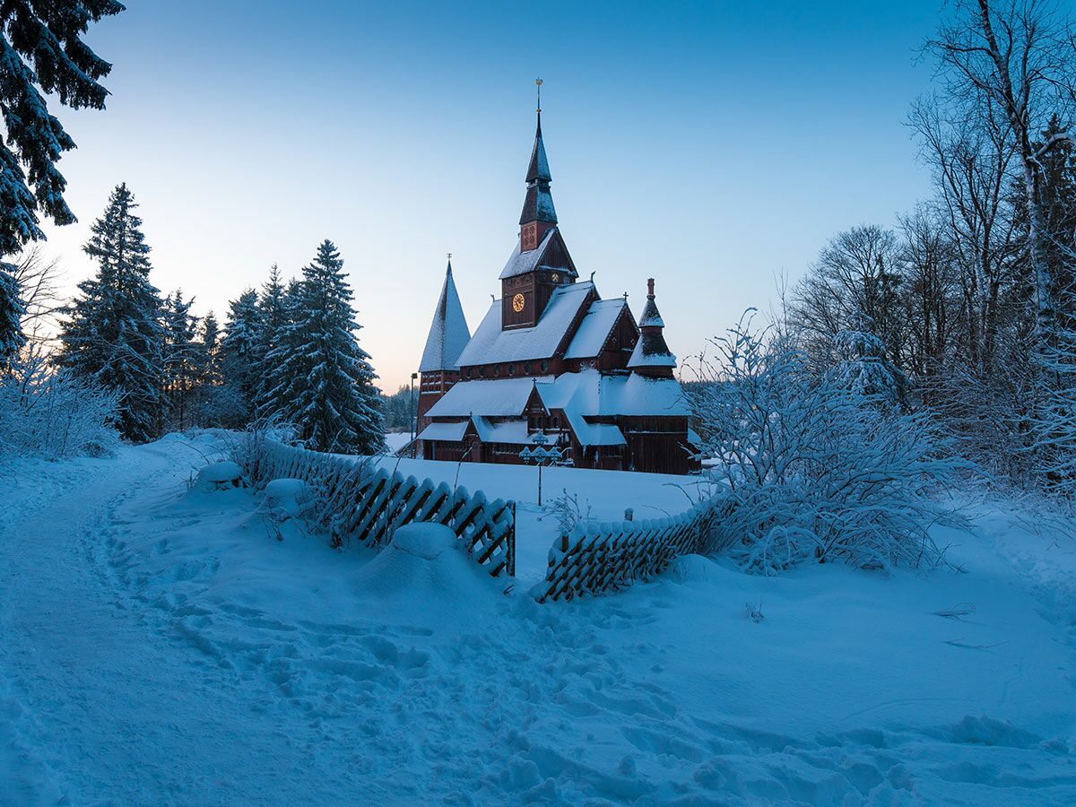 Die Hahnenklee Stabkirche im Winter, bedeckt mit Schnee, umgeben von verschneiten Bäumen und einem Pfad in der Dämmerung.