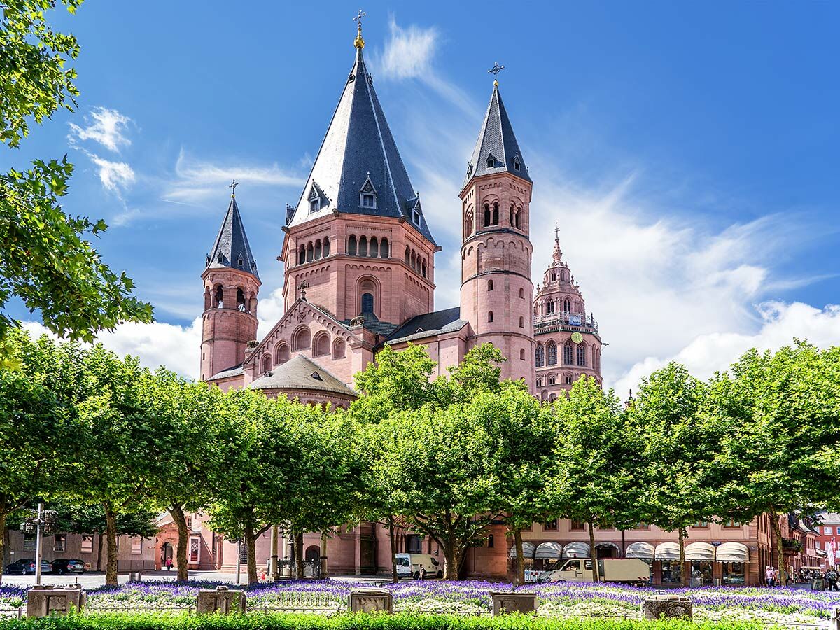 Der Mainzer Dom an einem sonnigen Tag mit blauem Himmel, umgeben von grünen Bäumen und blühenden Blumen auf dem Domplatz.