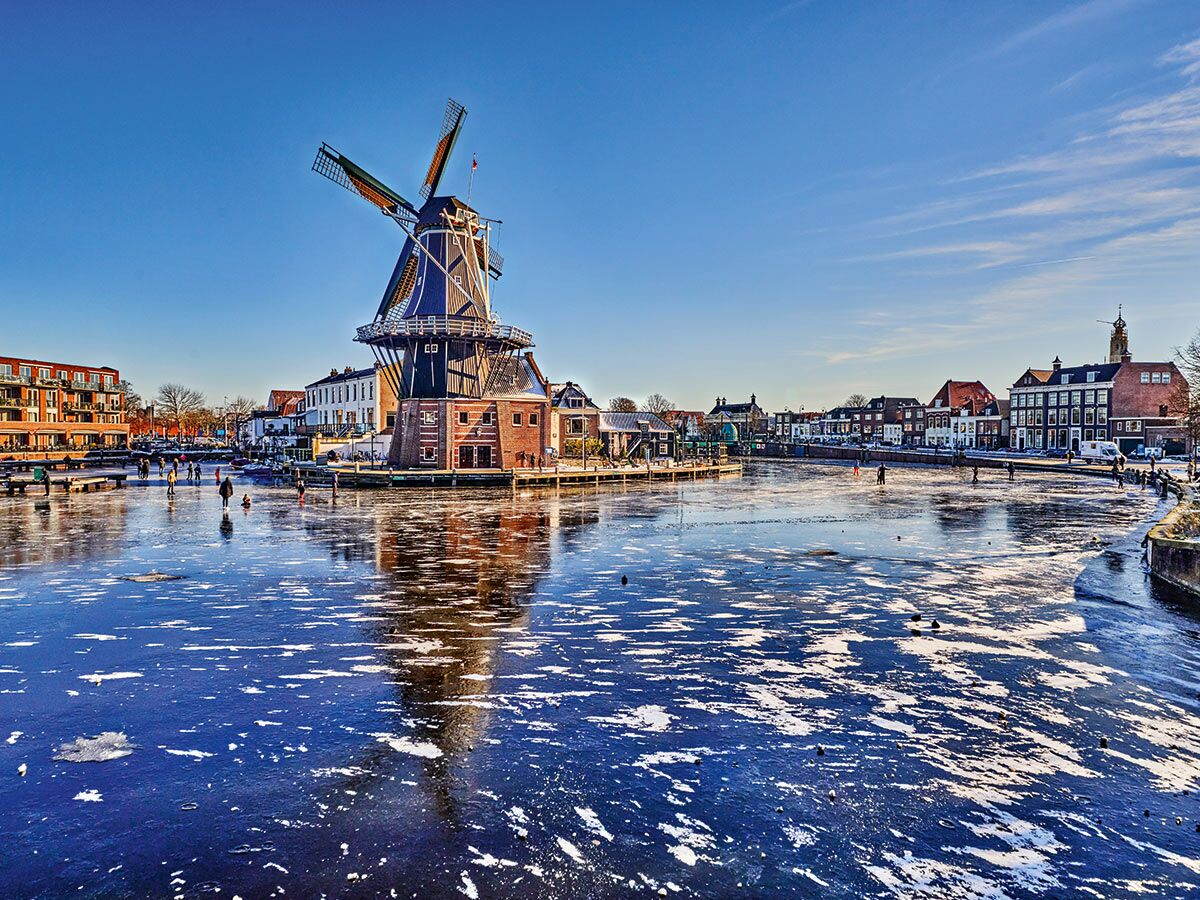 Historische Windmühle De Adriaan in Haarlem am gefrorenen Spaarne-Kanal mit Schlittschuhläufern an einem sonnigen Wintertag.