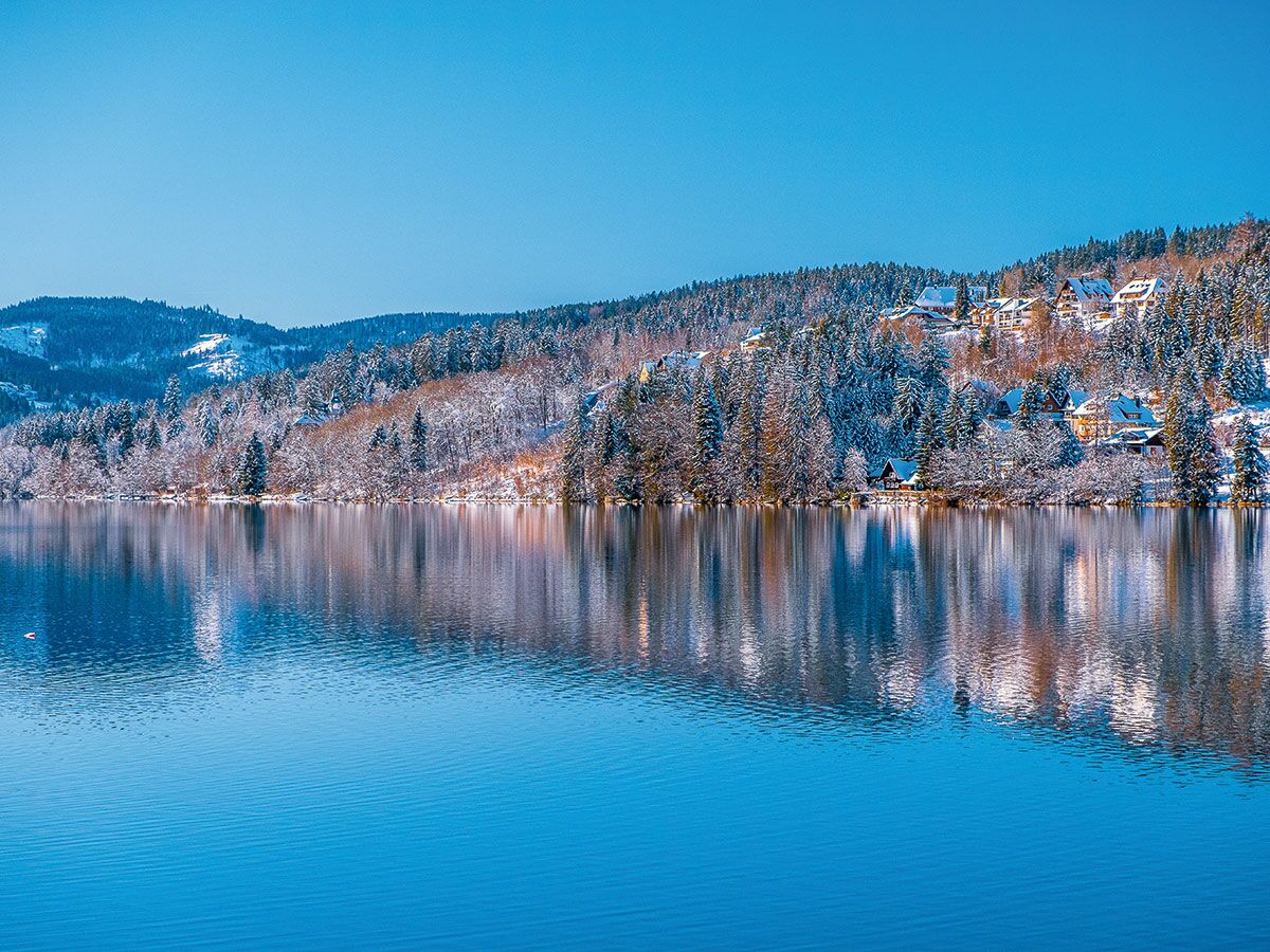 Ruhiger See mit spiegelndem Himmel und verschneiten Wäldern sowie Häusern an Berghängen unter blauem Himmel.