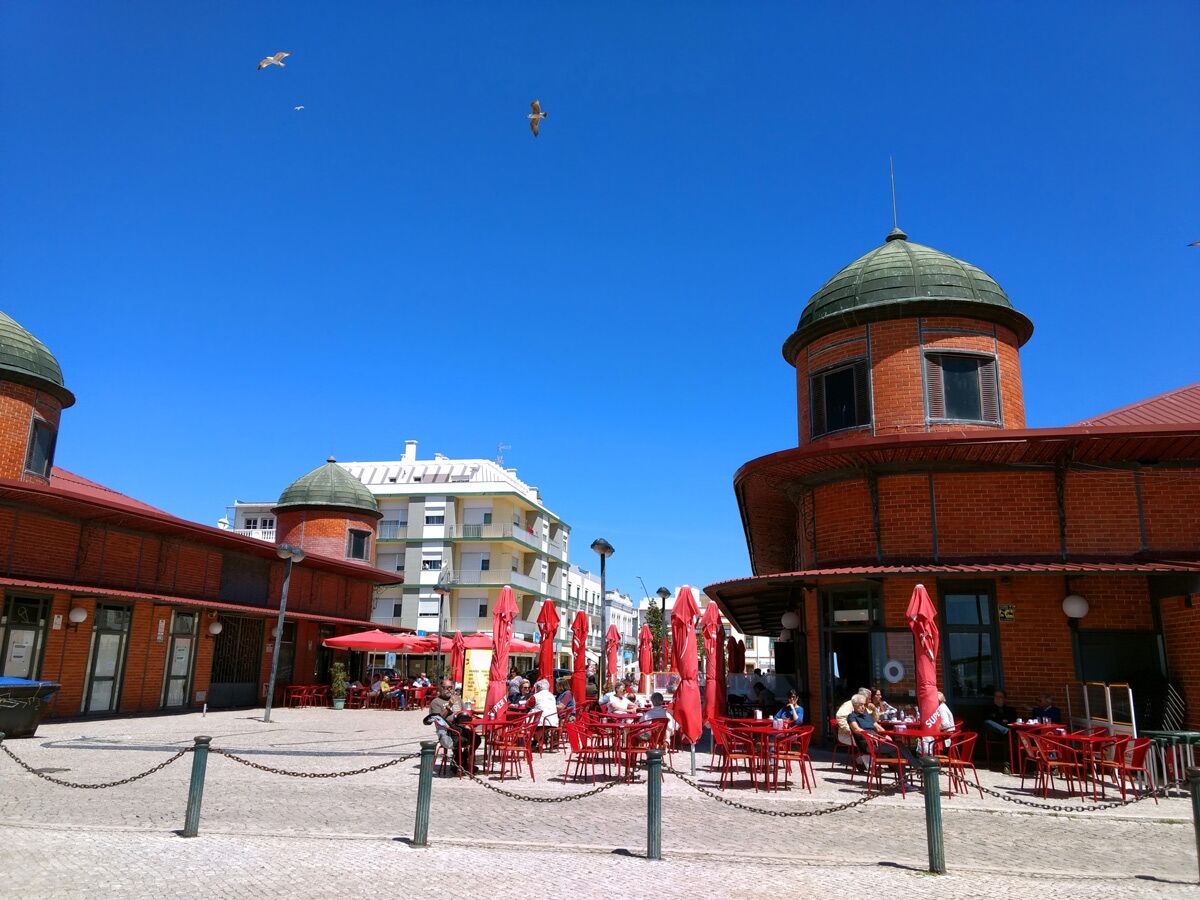 Menschen sitzen bei Sonnenschein im Außenbereich eines Cafés an einer historischen Markthalle mit Kuppeln unter roten Sonnenschirmen.