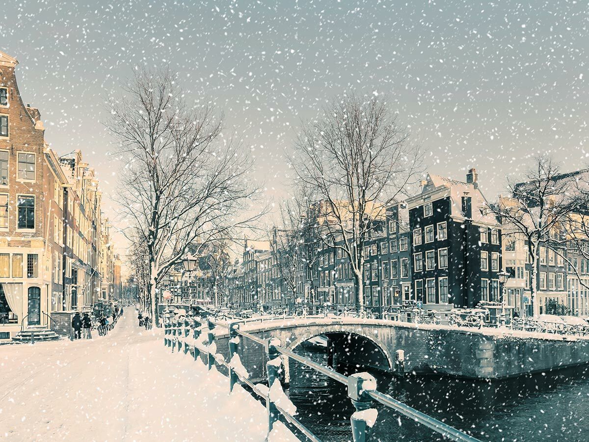 Verschneites Amsterdam im Winter: Kanal, Brücke, historische Häuser und Bäume unter fallenden Schneeflocken. Menschen spazieren.