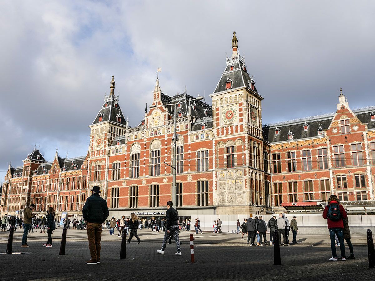 Historischer Hauptbahnhof Amsterdam mit rotem Backstein, Türmen und Uhren, belebter Vorplatz mit Menschen bei Tag.