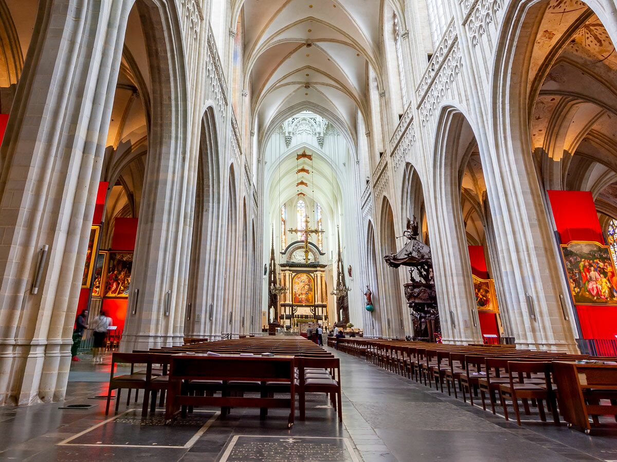 Innenansicht einer gotischen Kathedrale mit hohen Säulen, Gewölben, Kirchenbänken und roten Wandteppichen mit religiösen Gemälden.
