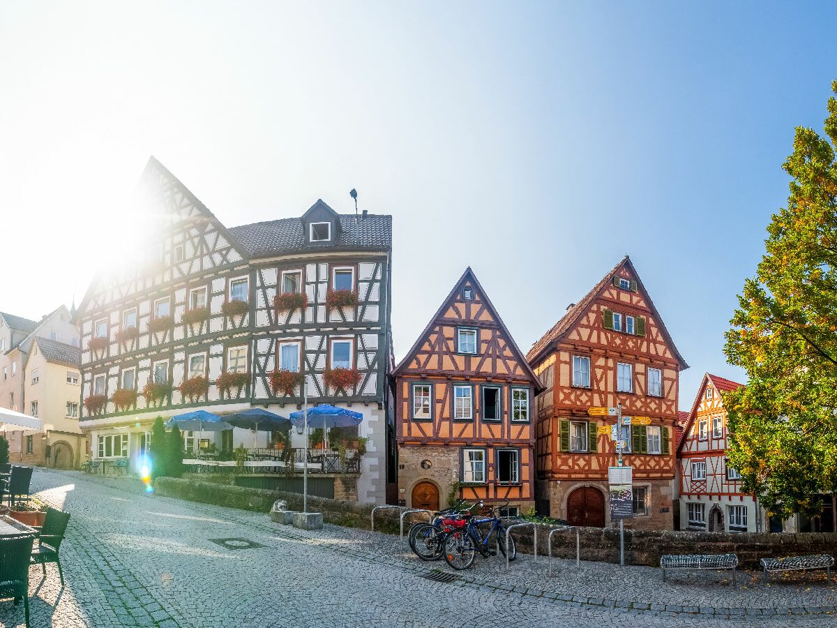 Historische Fachwerkhäuser an sonniger Kopfsteinpflastergasse in deutscher Altstadt mit Cafétischen, Fahrrädern und blauem Himmel.