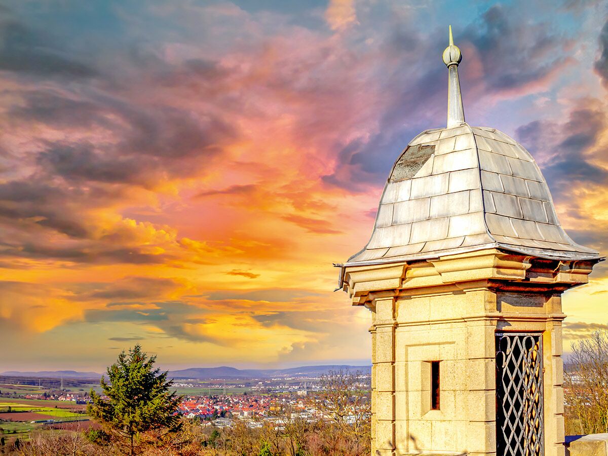 Alter steinerner Wachturm mit Metallkuppel vor dramatischem Sonnenuntergangshimmel über einem Dorf und Tal.