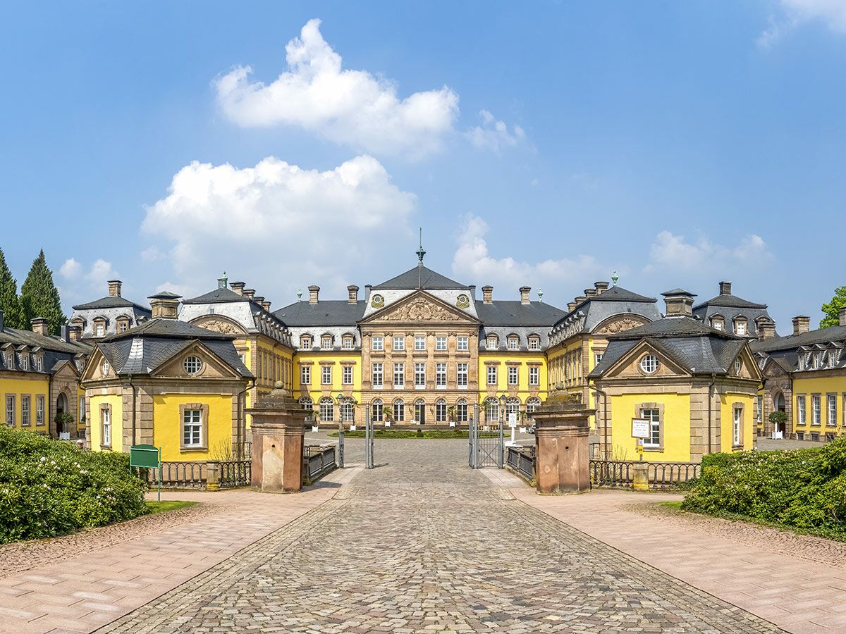 Großes, symmetrisches Barockschloss mit gelber Fassade, grauen Dächern und einem gepflasterten Vorplatz unter blauem Himmel.