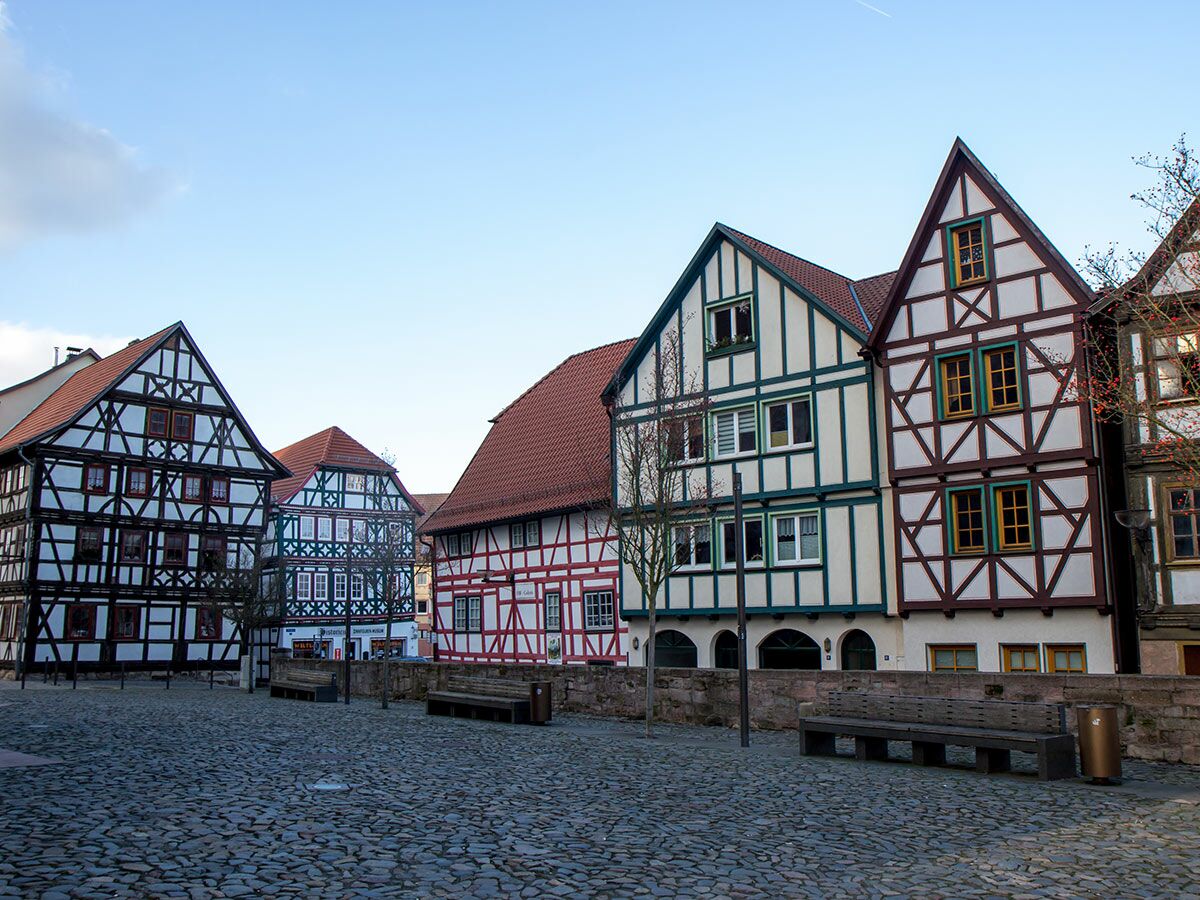Blick auf einen malerischen Platz mit traditionellen deutschen Fachwerkhäusern, Holzbänken und Kopfsteinpflaster unter blauem Himmel.
