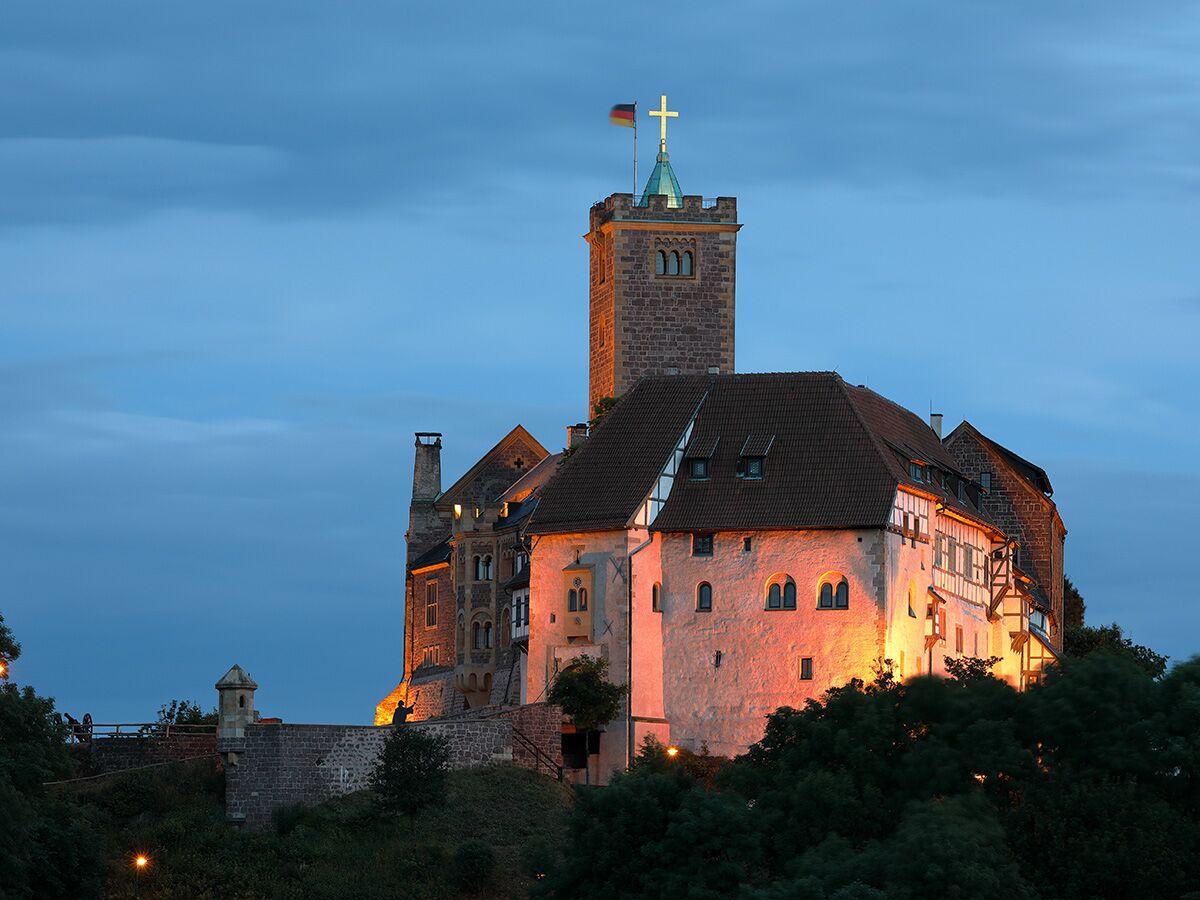 Historisches Wartburg Schloss in Thüringen bei Abenddämmerung, beleuchtet mit warmem Licht unter blauem Himmel und deutscher Flagge.