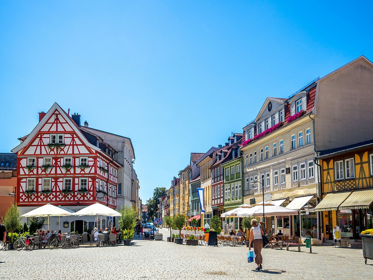 Belebte Altstadt-Gasse, Deutschland, mit farbenfrohen Fachwerkhäusern, Straßencafés, Menschen und Fahrrädern an sonnigem Tag.