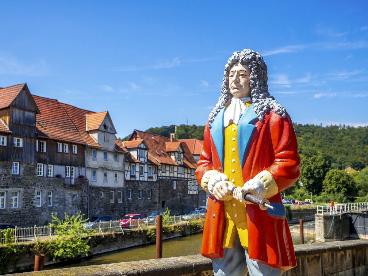Bunte Statue einer historischen Figur mit Perücke und rotem Mantel vor alten Häusern und Fluss unter blauem Himmel.