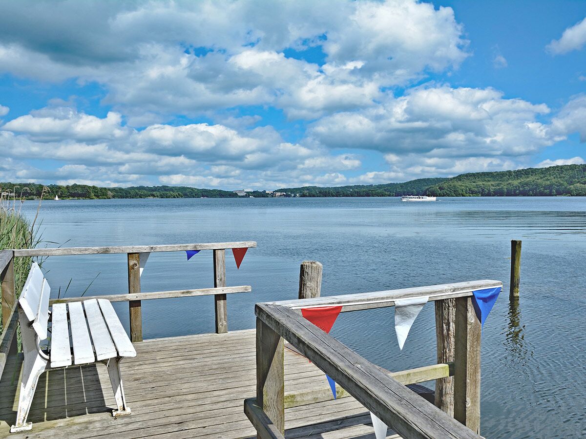 Blick von einem Holzsteg mit weißer Bank und bunten Wimpeln auf einen ruhigen See mit bewaldeten Ufern und Booten.