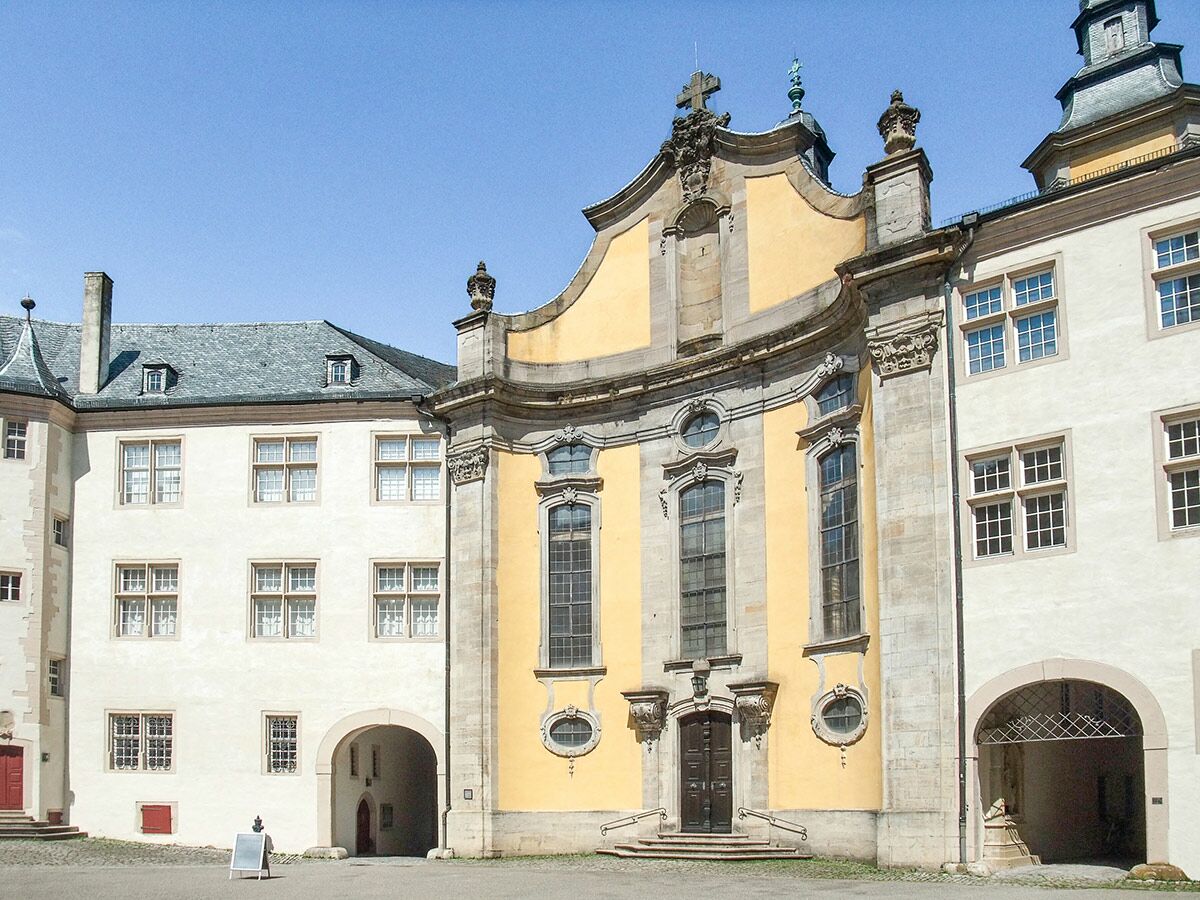 Barockschloss mit gelber Kirchenfassade und hellen Gebäuden im Innenhof unter blauem Himmel.