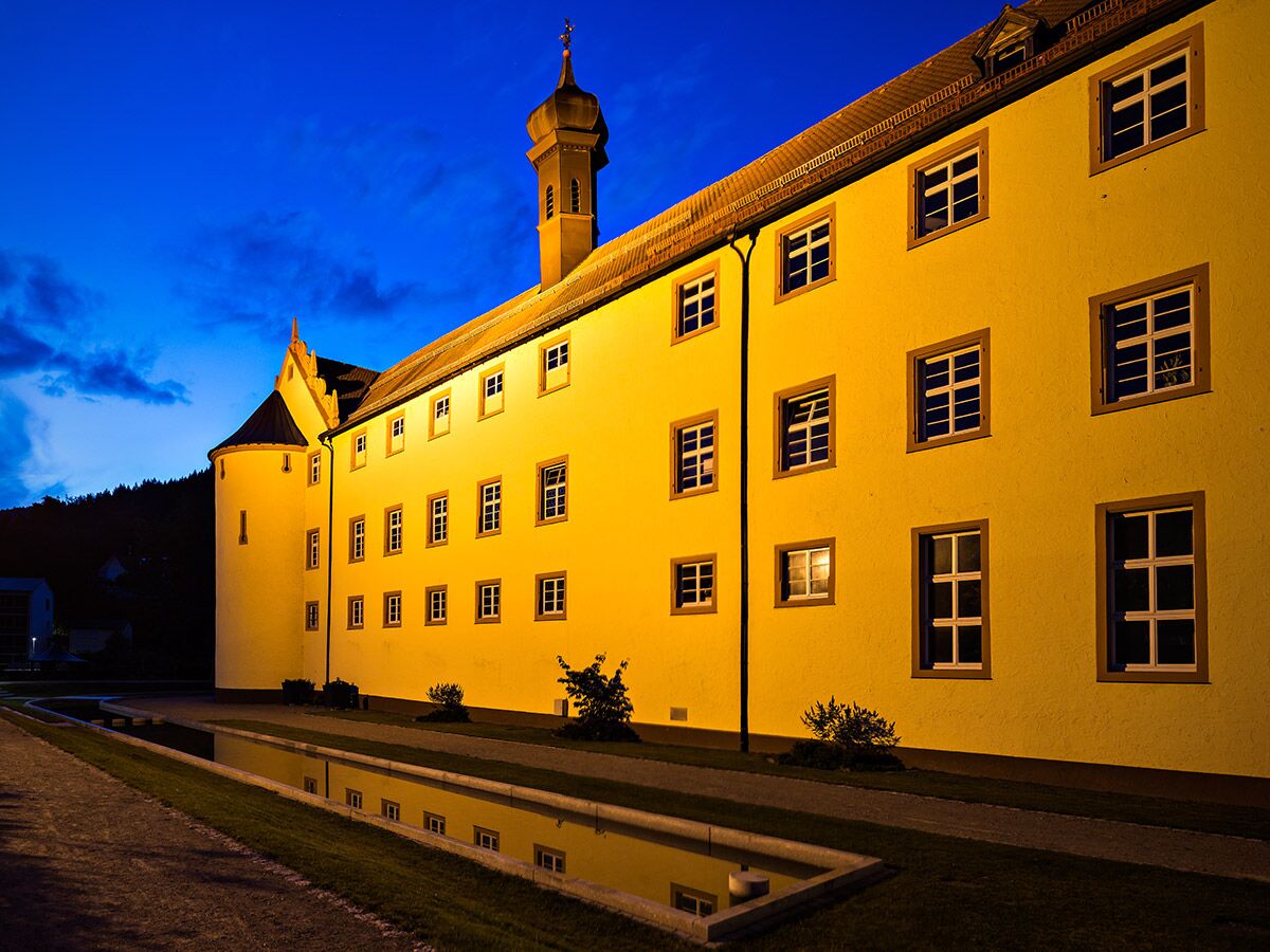 Nachtaufnahme eines hell beleuchteten gelben Klostergebäudes mit Turm und Fensterreflexion im Wasserbecken.