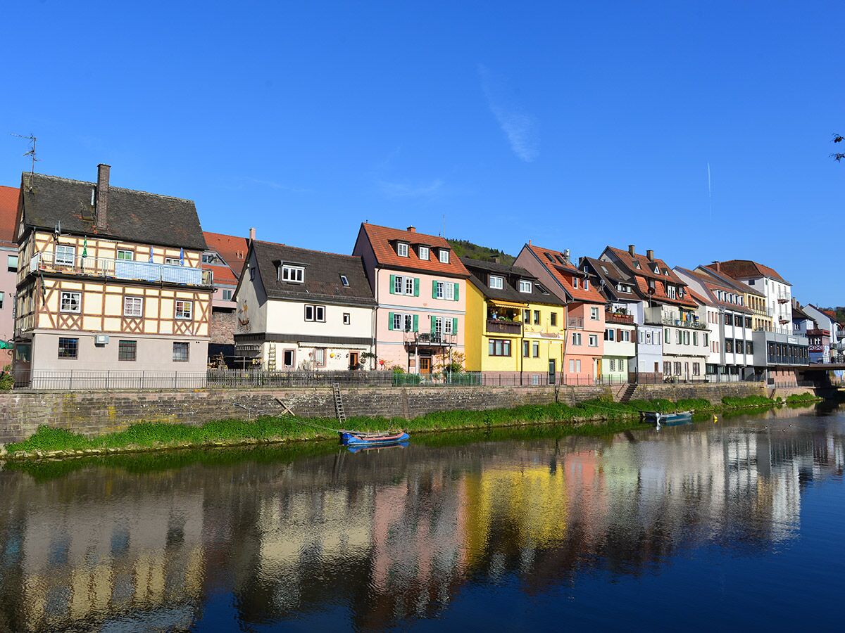 Reihe bunter Häuser am Flussufer mit Spiegelungen im Wasser unter blauem Himmel in einer malerischen deutschen Stadt.