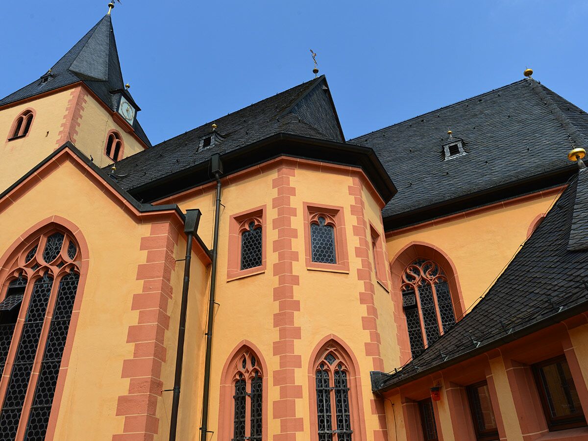 Detailansicht einer historischen Kirche mit orange-roter Fassade, Spitzbogenfenstern, dunklem Schieferdach und hohem Glockenturm unter blauem Himmel.