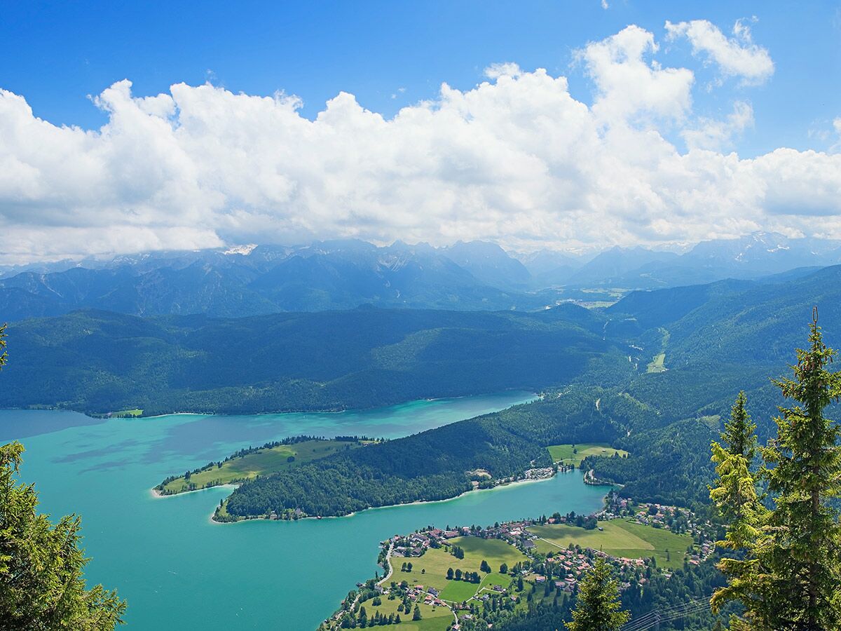 Blick auf den türkisblauen Walchensee, umgeben von grünen Wäldern und bayerischen Alpen, mit einem Dorf am Ufer unter blauem Himmel.