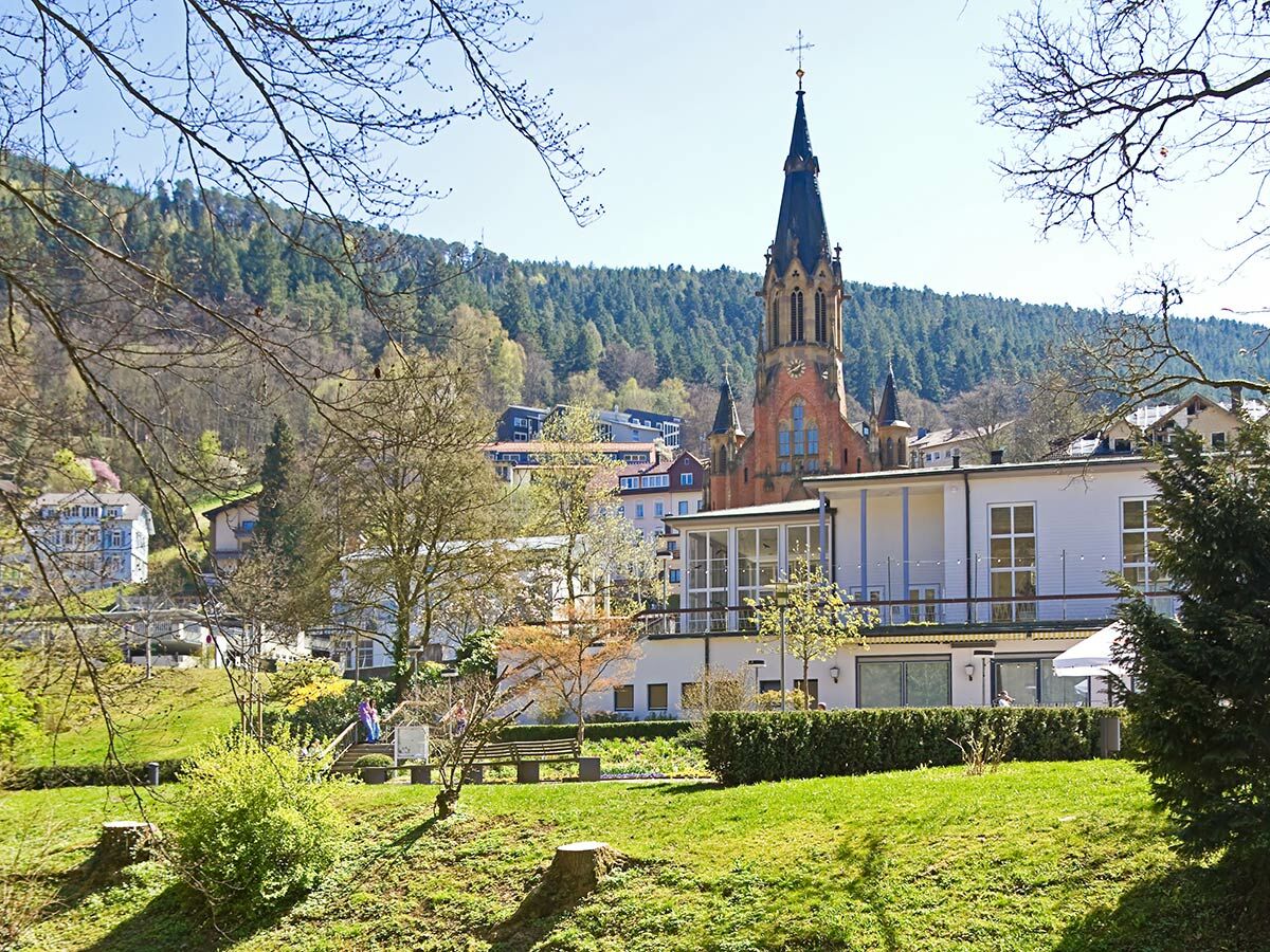 Panoramablick auf die Stadtkirche Bad Wildbad mit grüner Parkanlage und bewaldeten Hügeln im Frühling unter blauem Himmel.