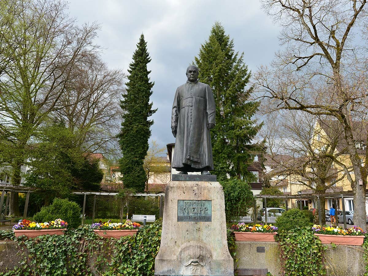 Statue von Sebastian Kneipp in einem Park, umgeben von Bäumen, Blumen und Efeu unter bewölktem Himmel.