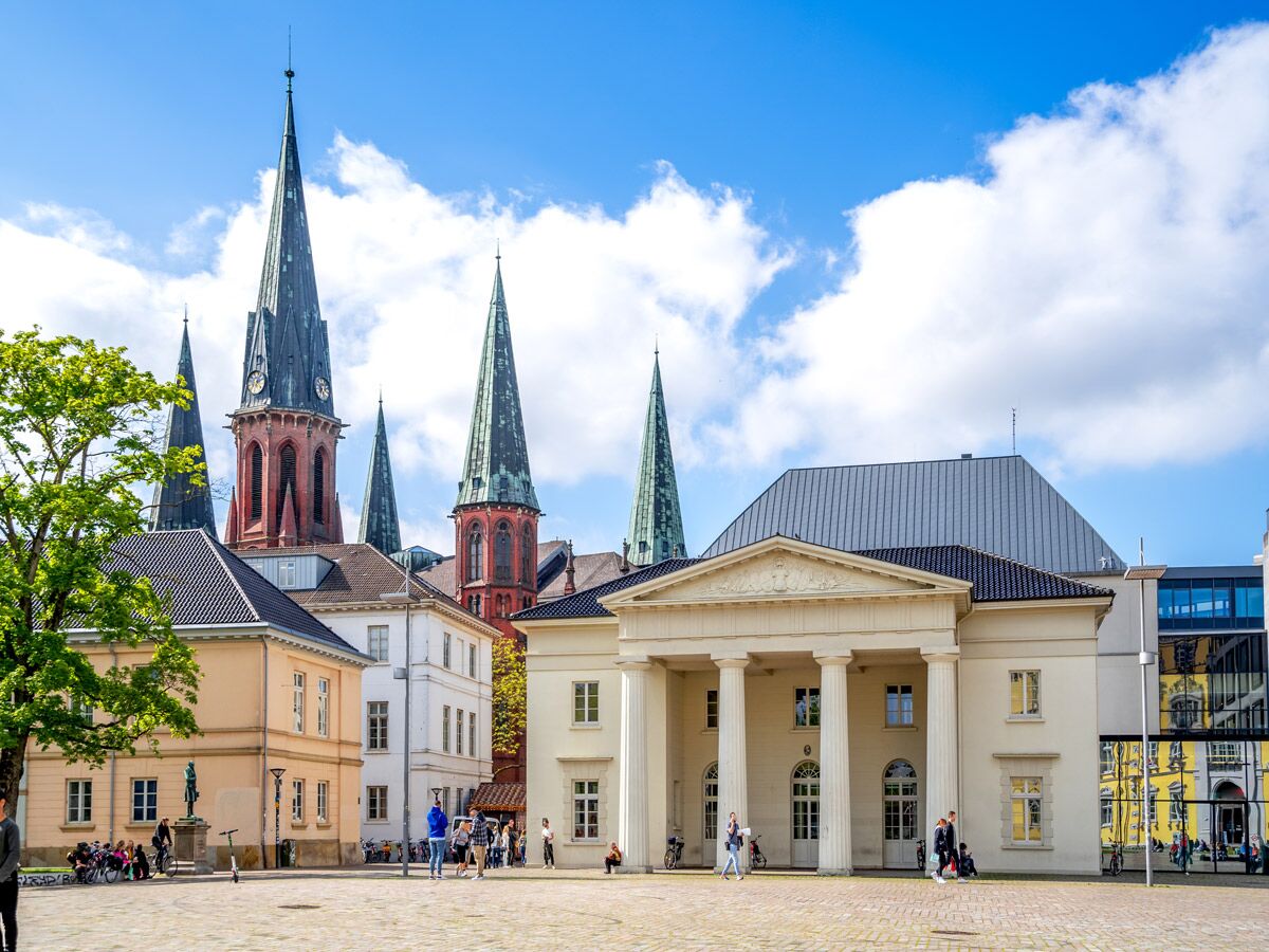 Marktplatz Oldenburg mit Staatstheater, Lamberti-Kirche und historischen Gebäuden unter blauem Himmel. Menschen spazieren.
