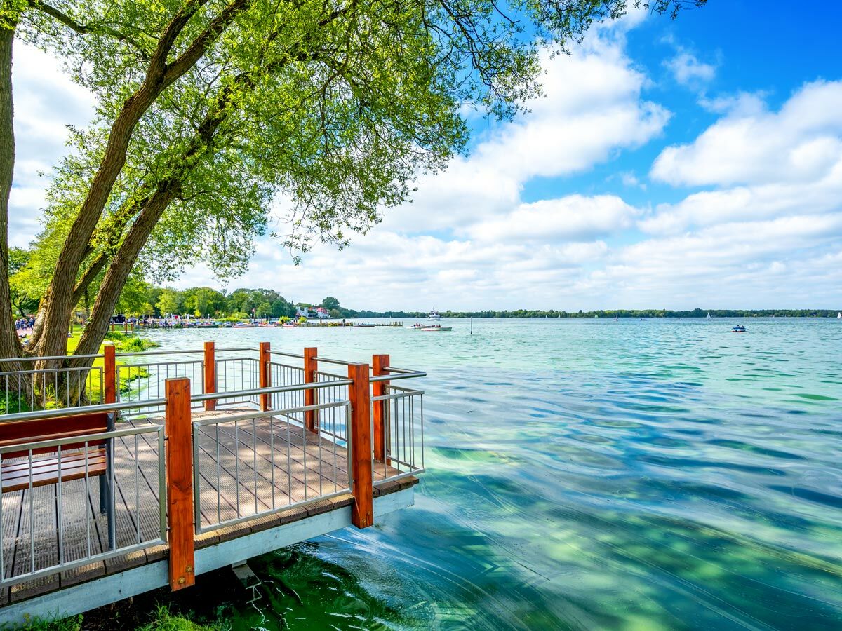 Idyllischer See mit Holzsteg, grünen Bäumen und blauem Himmel. Boote sind auf dem klaren Wasser sichtbar, Ufer in der Ferne.