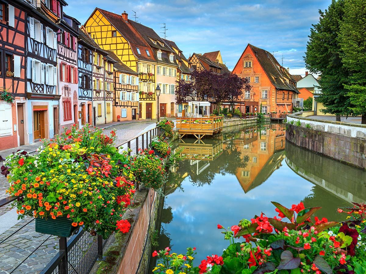 Bunte Fachwerkhäuser und üppige Blumenarrangements säumen einen malerischen Kanal in Colmar, Frankreich, mit Spiegelungen im Wasser.