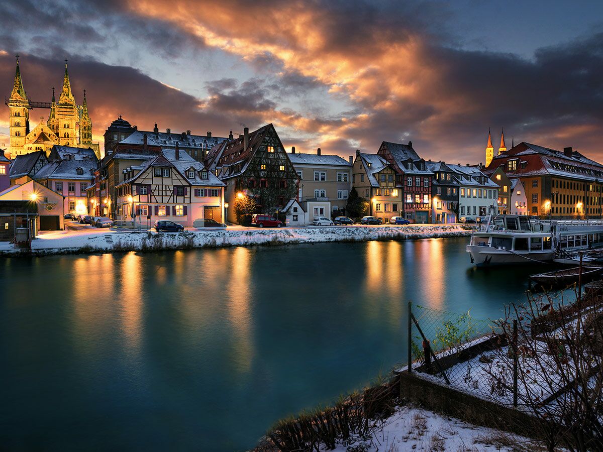 Panoramablick auf das verschneite Bamberg im Abendlicht. Historische Häuser und Boote spiegeln sich im Fluss wider, dramatischer Himmel.