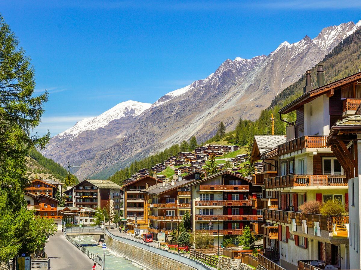 Panorama eines malerischen Schweizer Bergdorfs mit traditionellen Chalets, Fluss im Tal und schneebedeckten Alpen im Hintergrund.