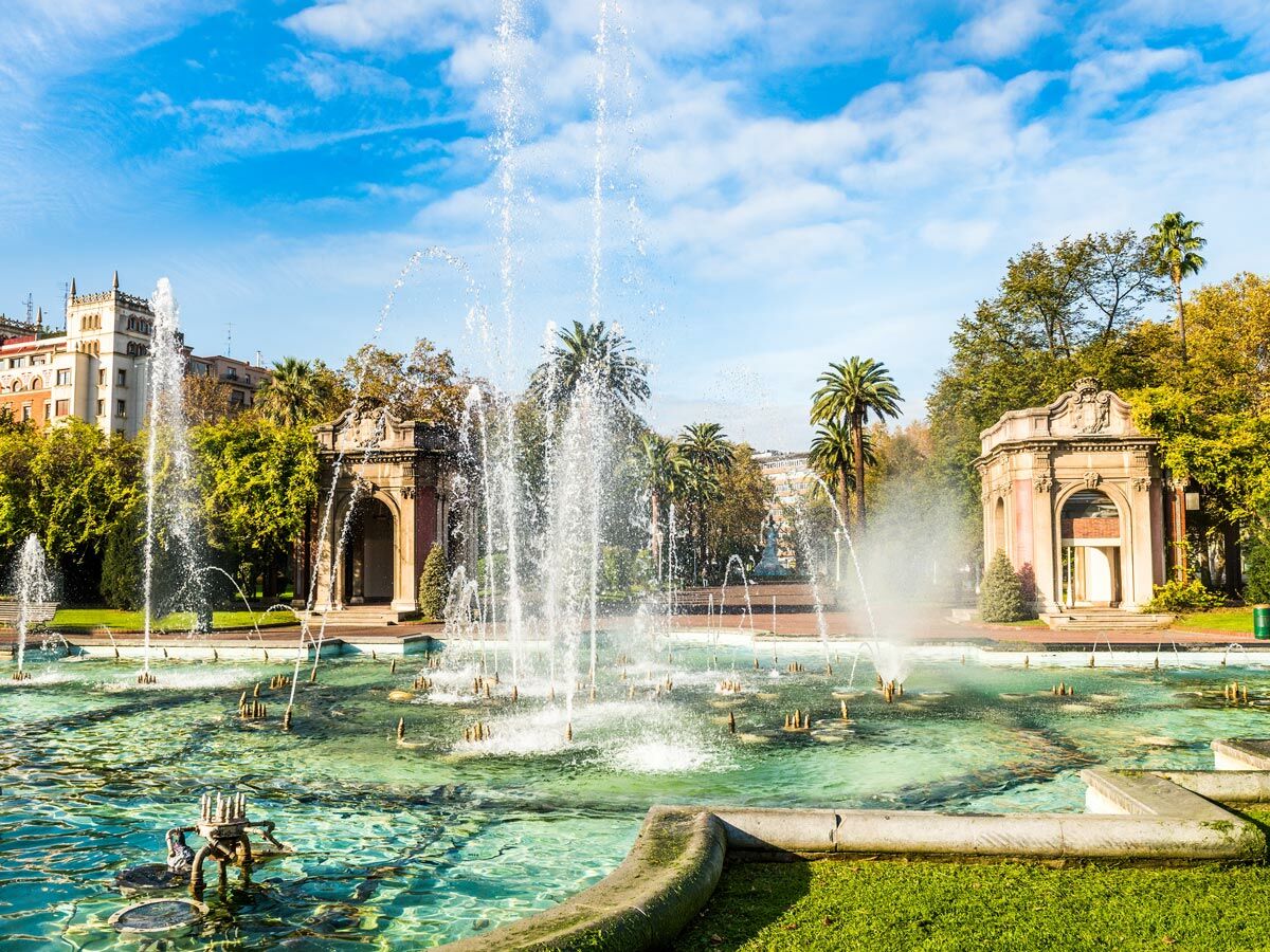Großer Springbrunnen mit vielen Wasserstrahlen in einem sonnigen, grünen Park. Im Hintergrund sind historische Gebäude und Palmen zu sehen.