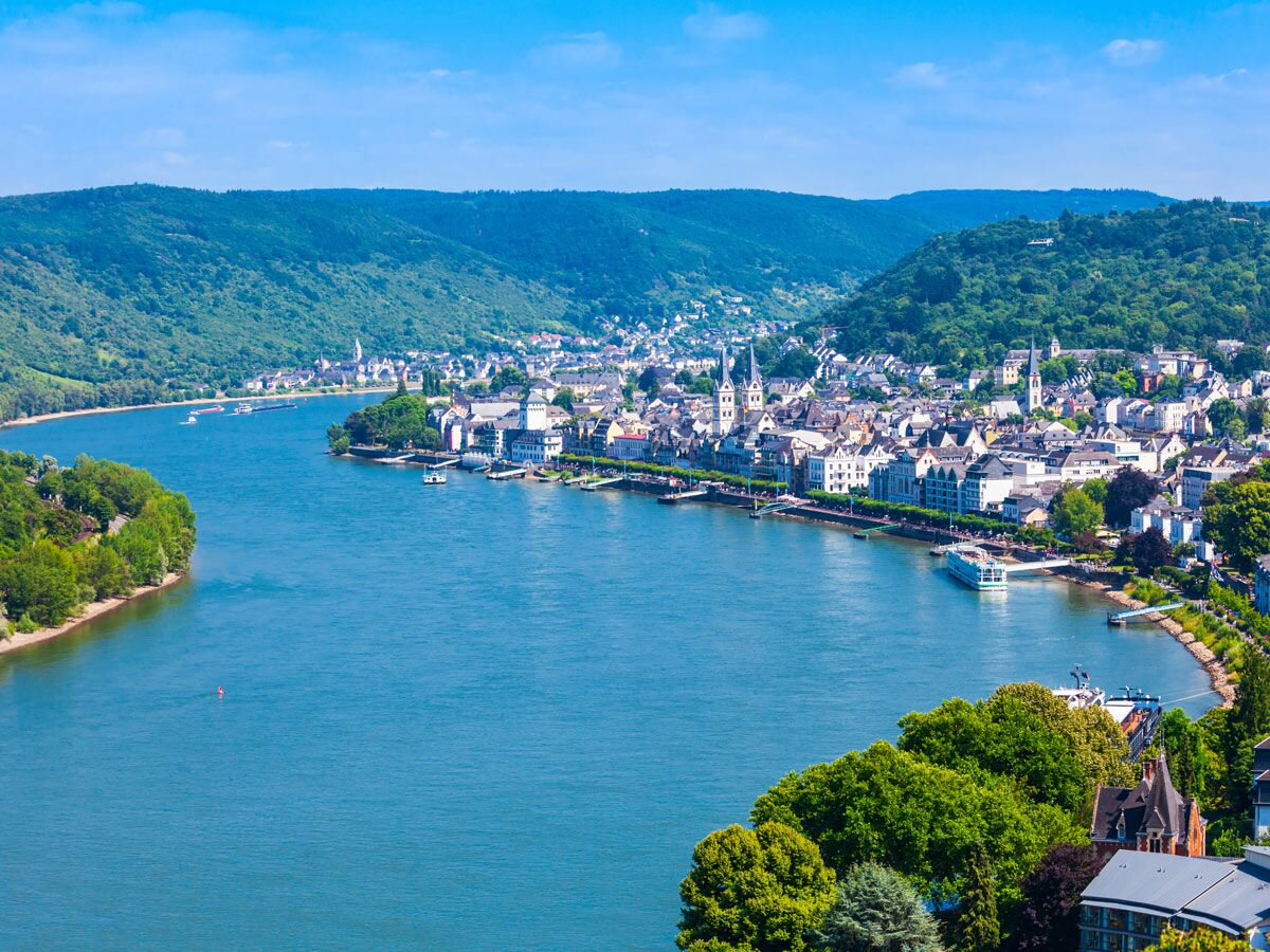 Panoramablick auf die Stadt Boppard am Rhein mit historischen Gebäuden, Schifffahrt und grünen Hügeln unter blauem Himmel.