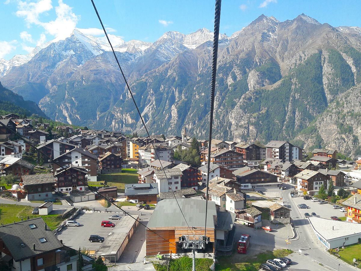 Malerische Ansicht eines traditionellen Schweizer Bergdorfs im Tal mit schneebedeckten Bergen, von einer Seilbahn aus gesehen.